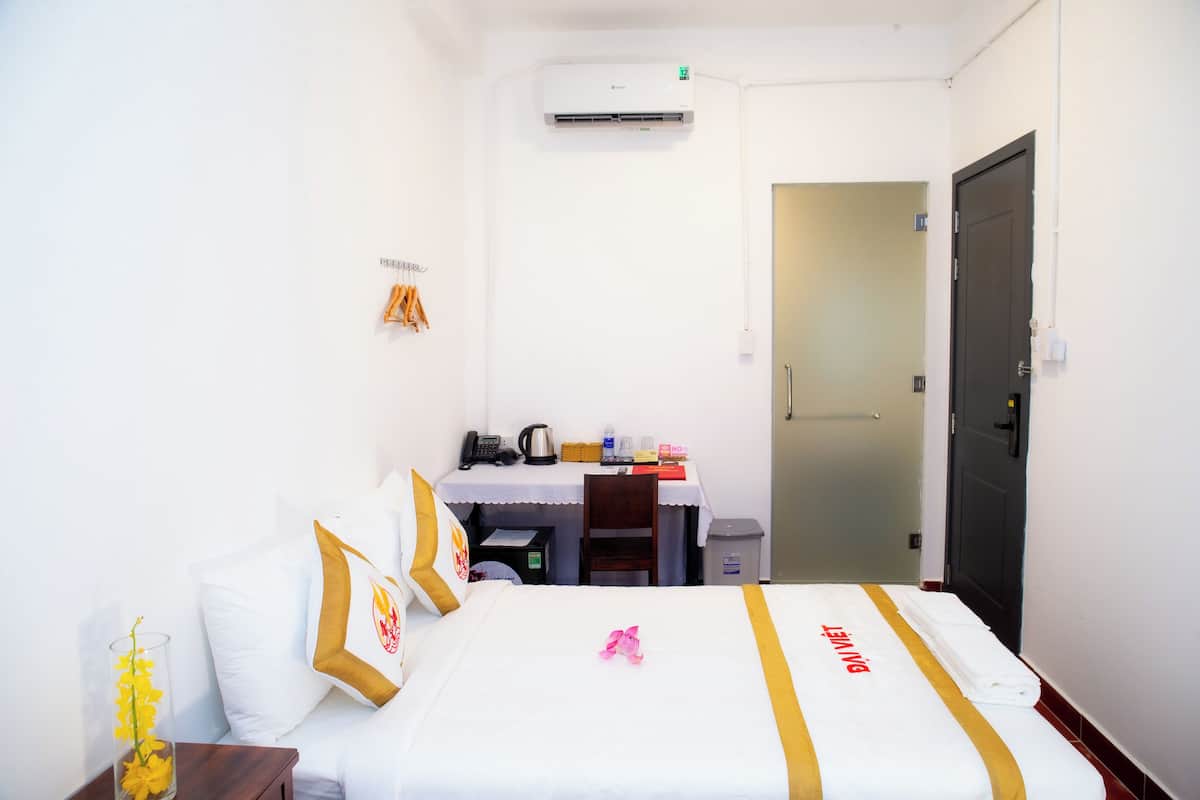 A hotel room with a bed, a desk, and a chair. There is a trash bin and a door.