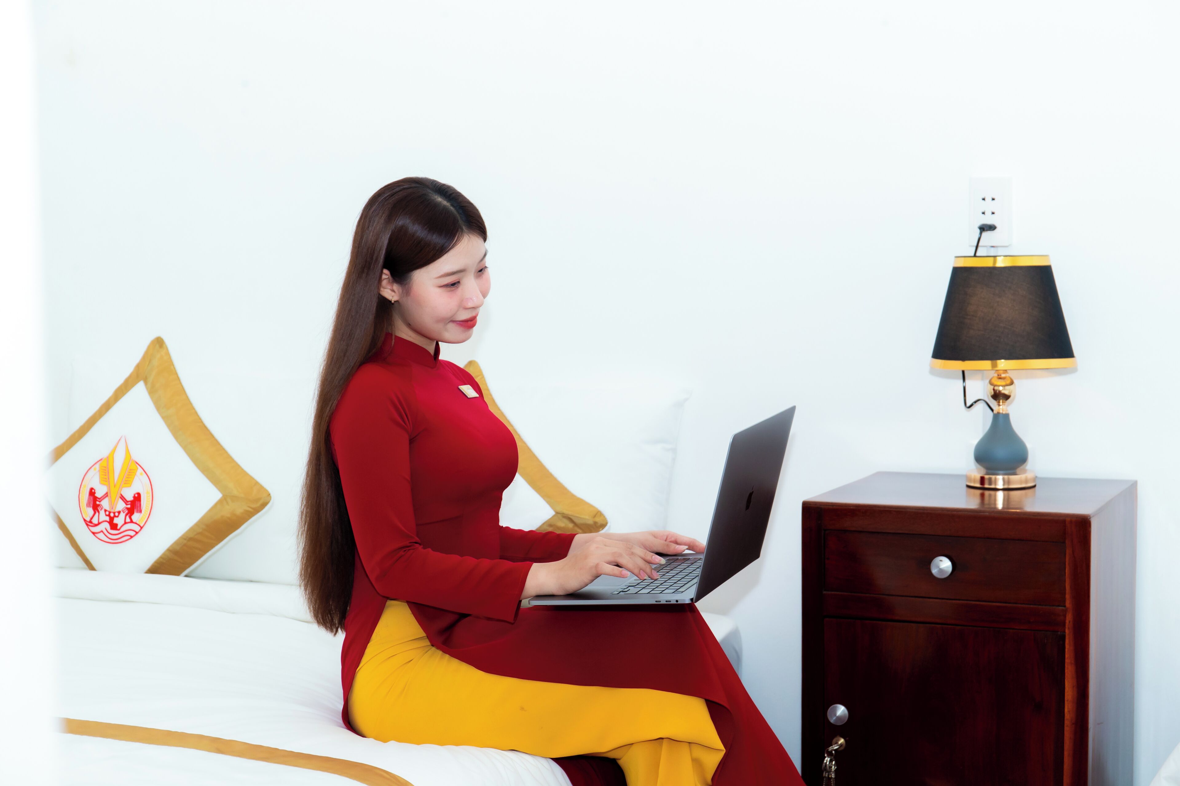 Une personne assise sur un lit, avec un ordinateur portable, une table de chevet avec une lampe et un mur décoré d’un coussin.