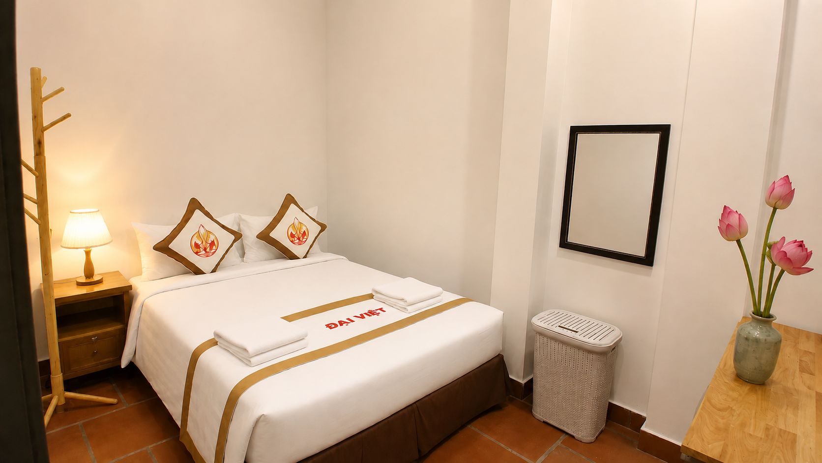A hotel room with a neatly made bed, a bedside lamp, a trash bin, a framed mirror, and a vase with flowers.