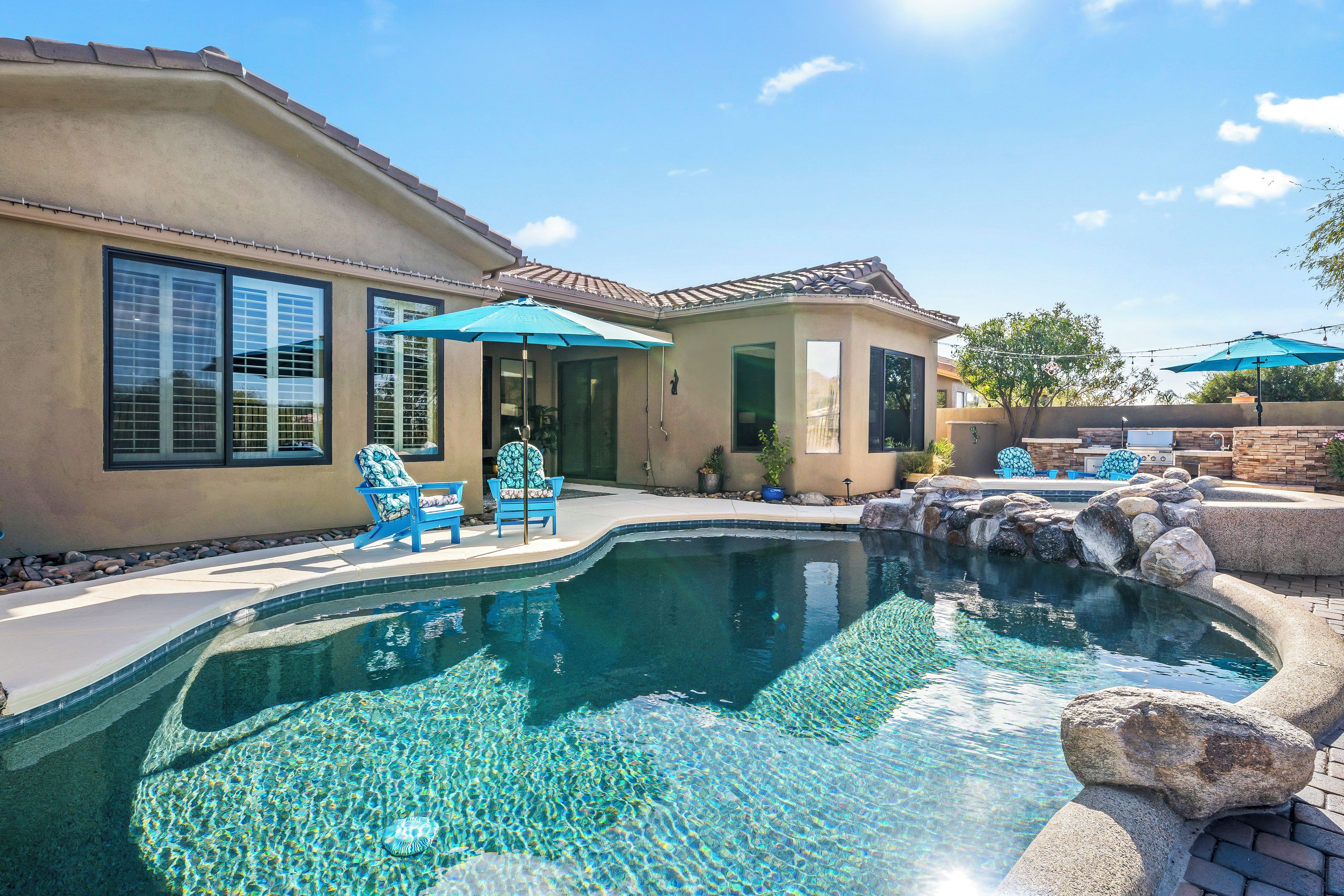A residential swimming pool with clear water, surrounded by a patio area with chairs and umbrellas.