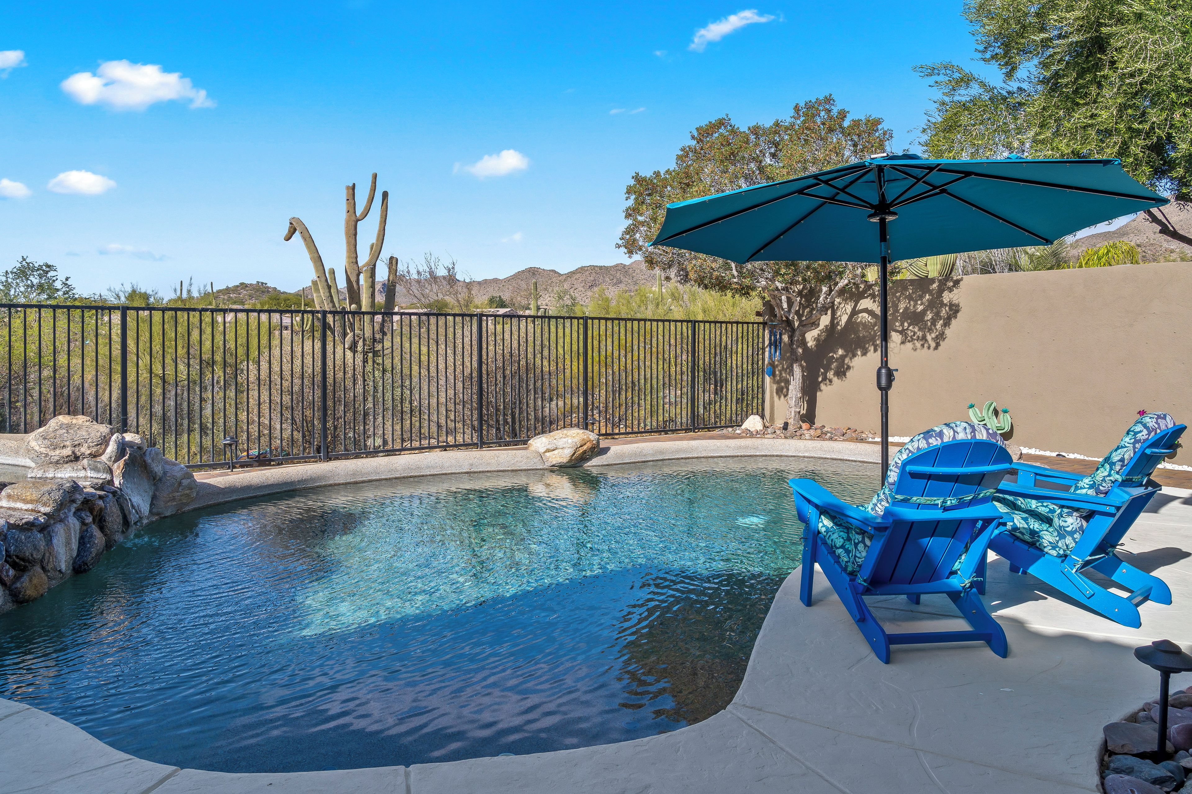 A swimming pool with a blue chair and umbrella, surrounded by a fence and desert vegetation.