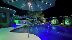 Zona de piscina con borde curvo, rodeada por una valla e iluminada con luces colgantes.