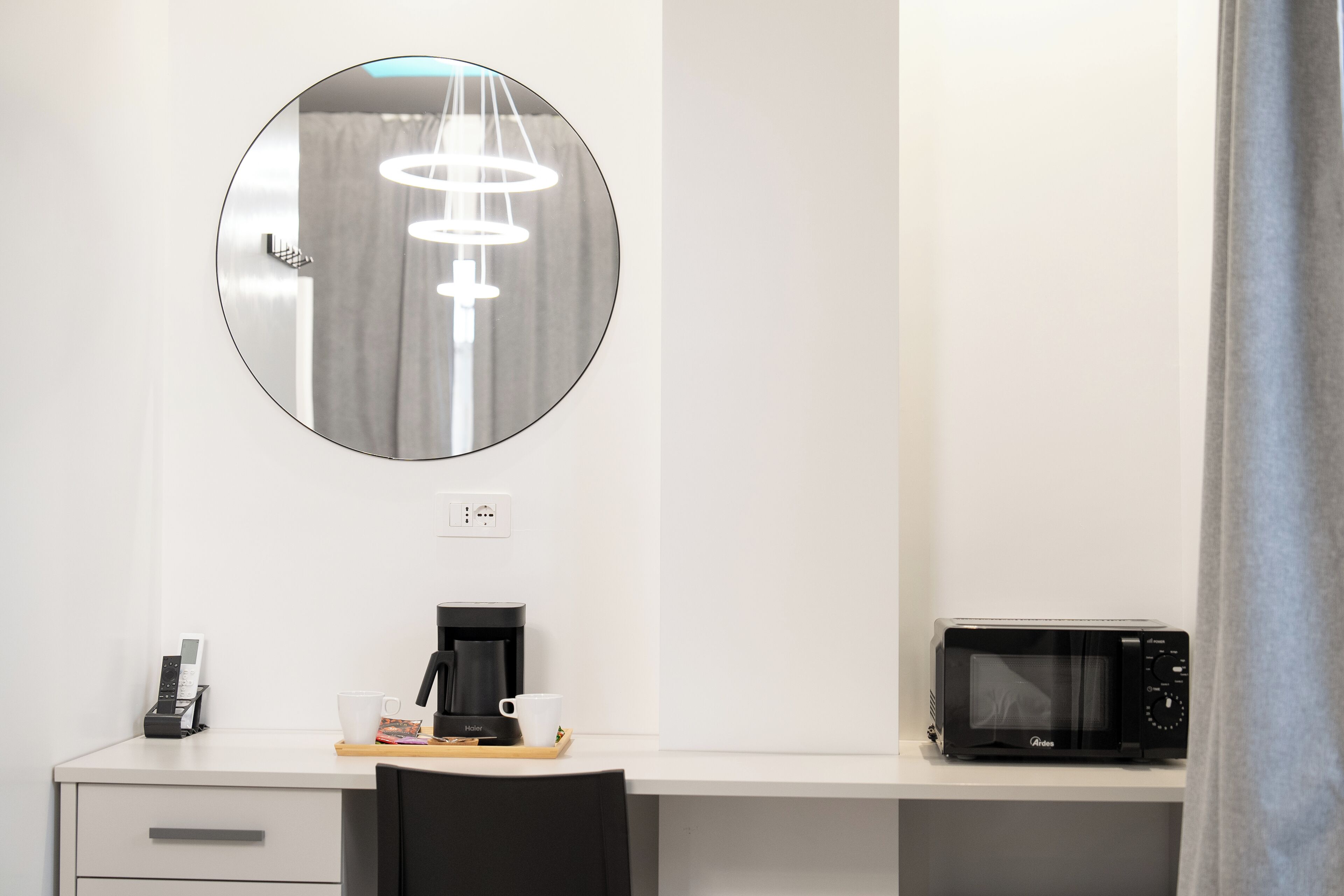 Une chambre d’hôtel moderne avec un miroir rond, une machine à café, un four à micro-ondes et une petite table sur laquelle se trouvent une tasse et un livre.