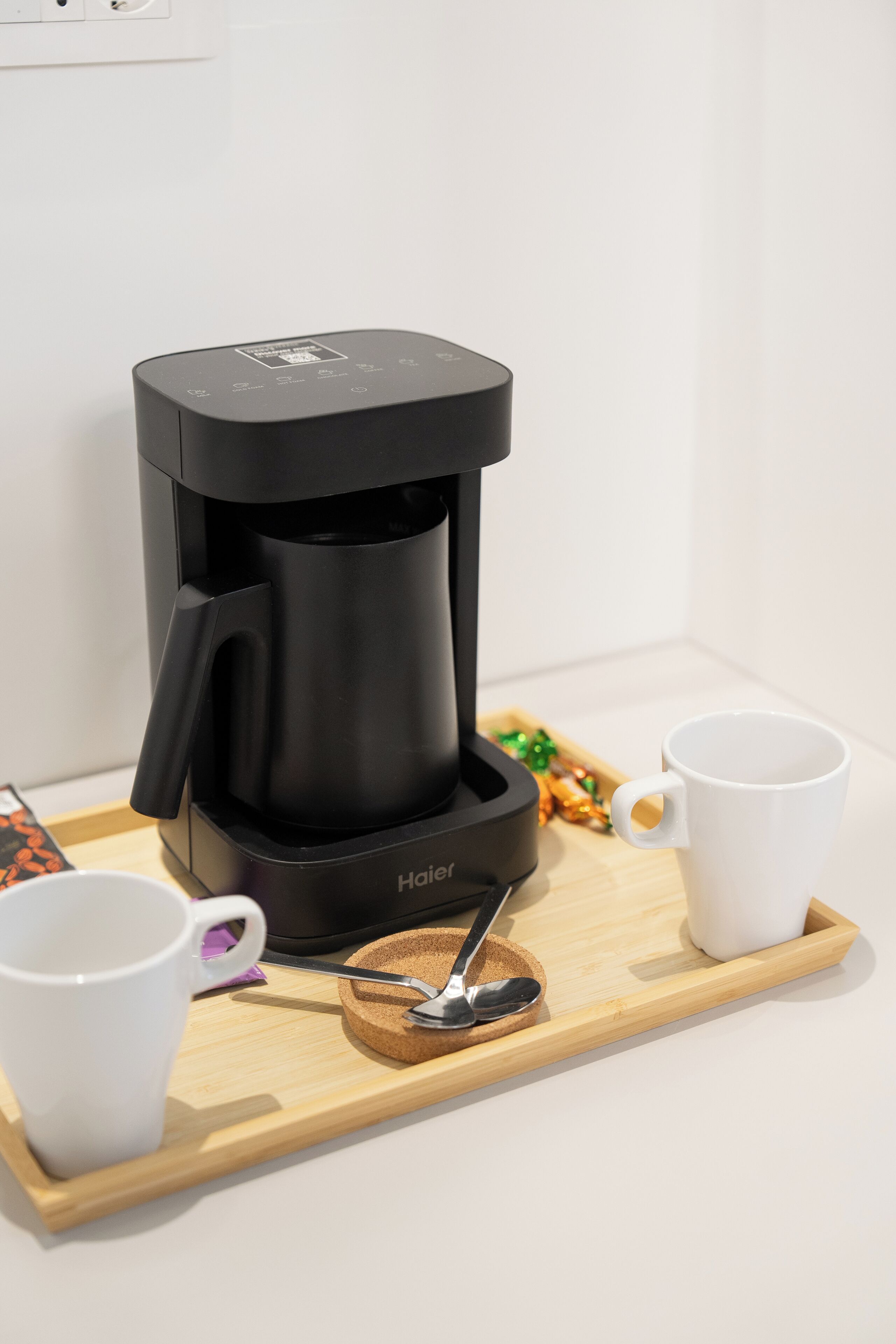 Eine Kaffeemaschine mit schwarzem Gehäuse auf einer Holztablett zusammen mit zwei weißen Tassen und einem Löffel.