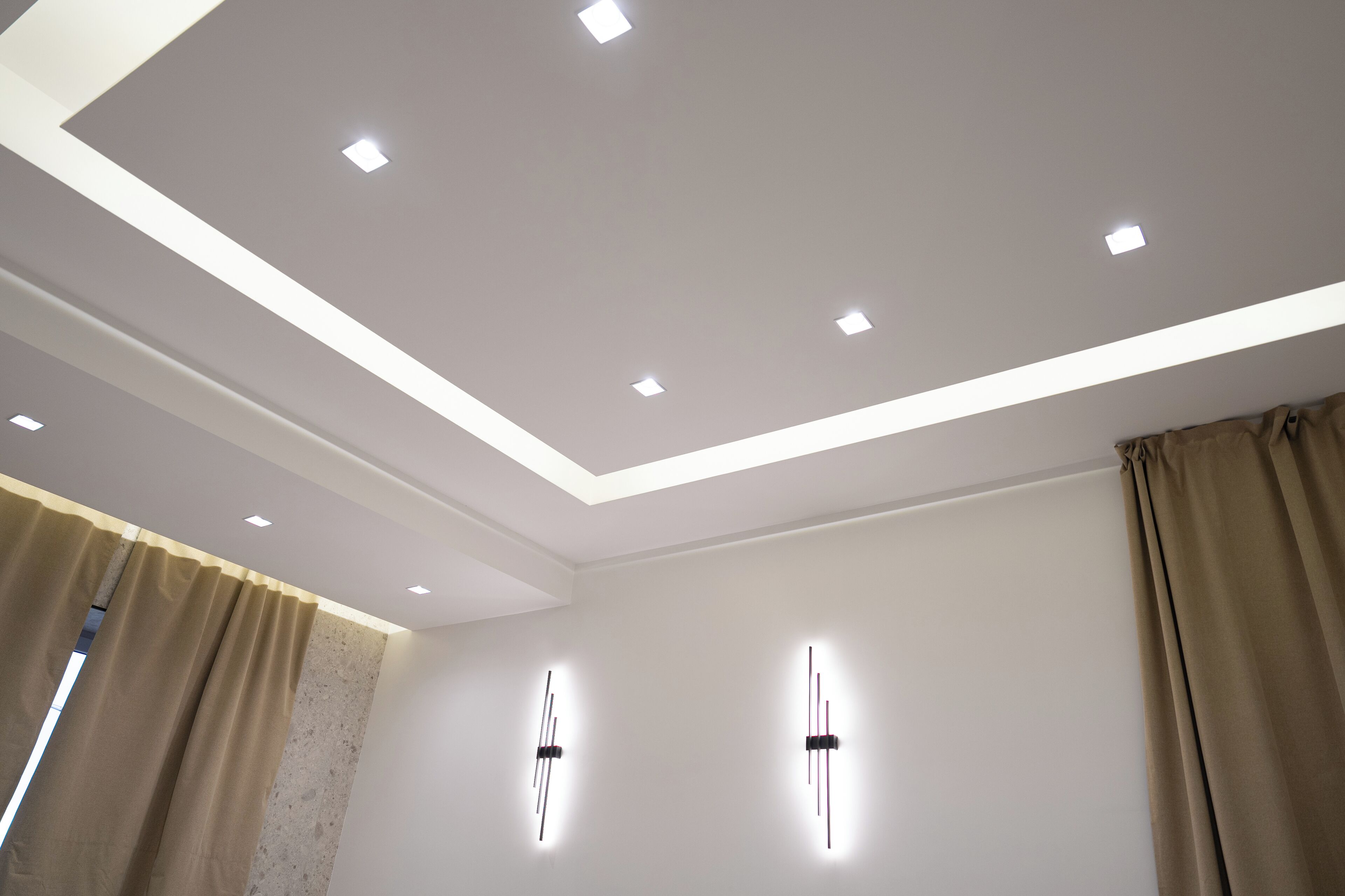 Una habitación moderna con iluminación empotrada en el techo, una pared lisa y un ventanal con cortinas.