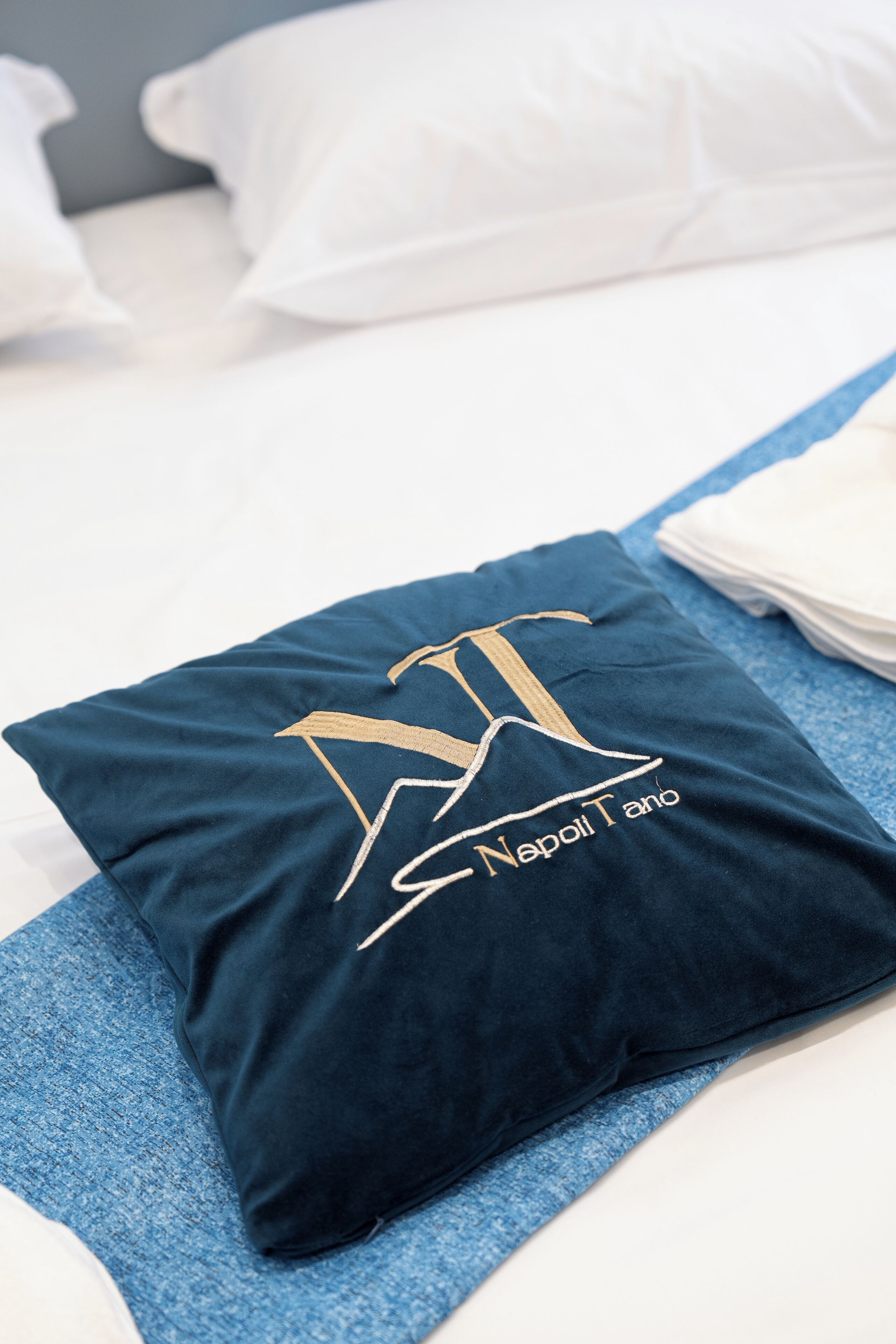 A neatly made bed with a dark blue pillow featuring a gold embroidered logo and text, and a stack of white pillows.