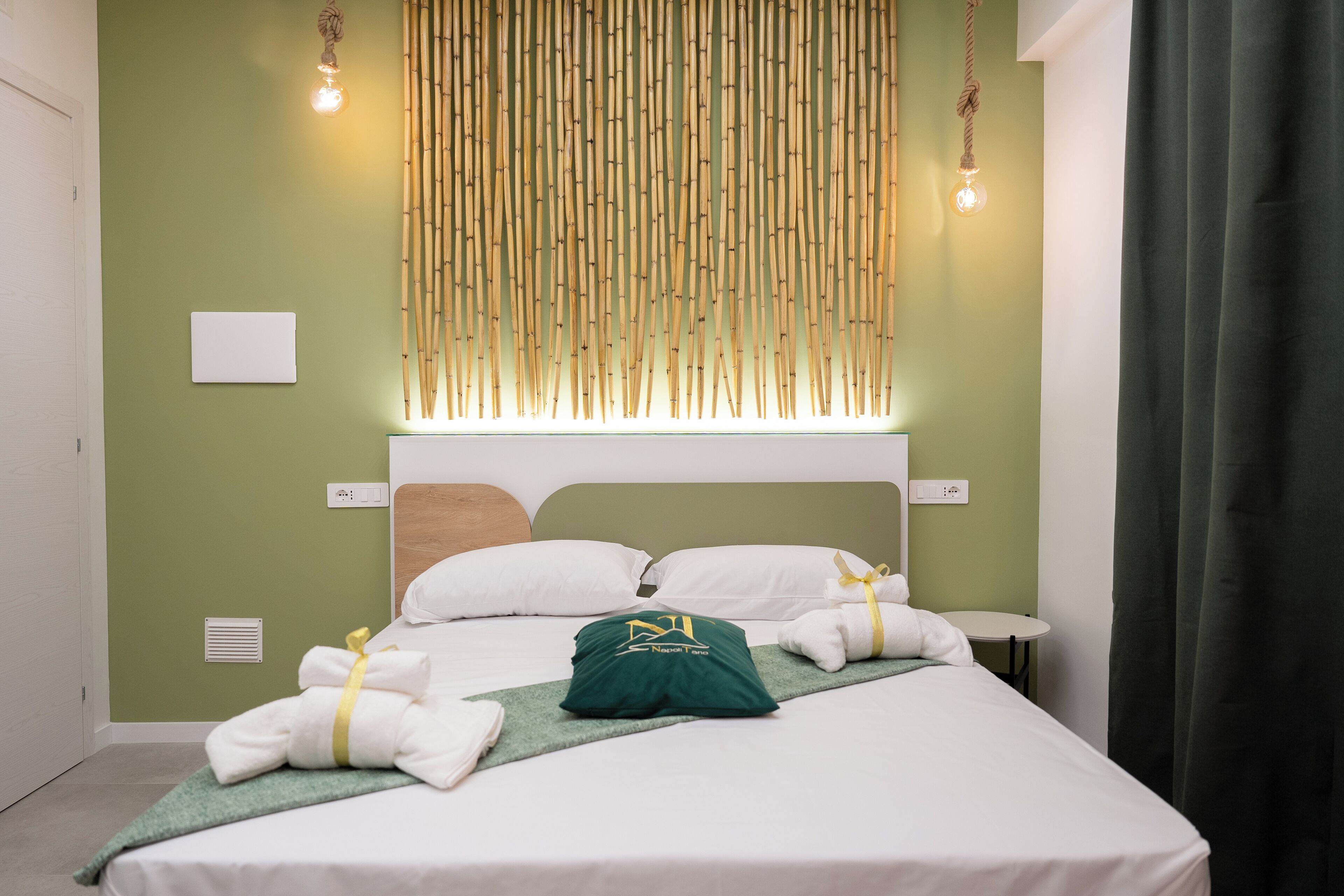A neatly made bed with white linens and a green towel, a bedside table with a lamp, and a wall-mounted light fixture.