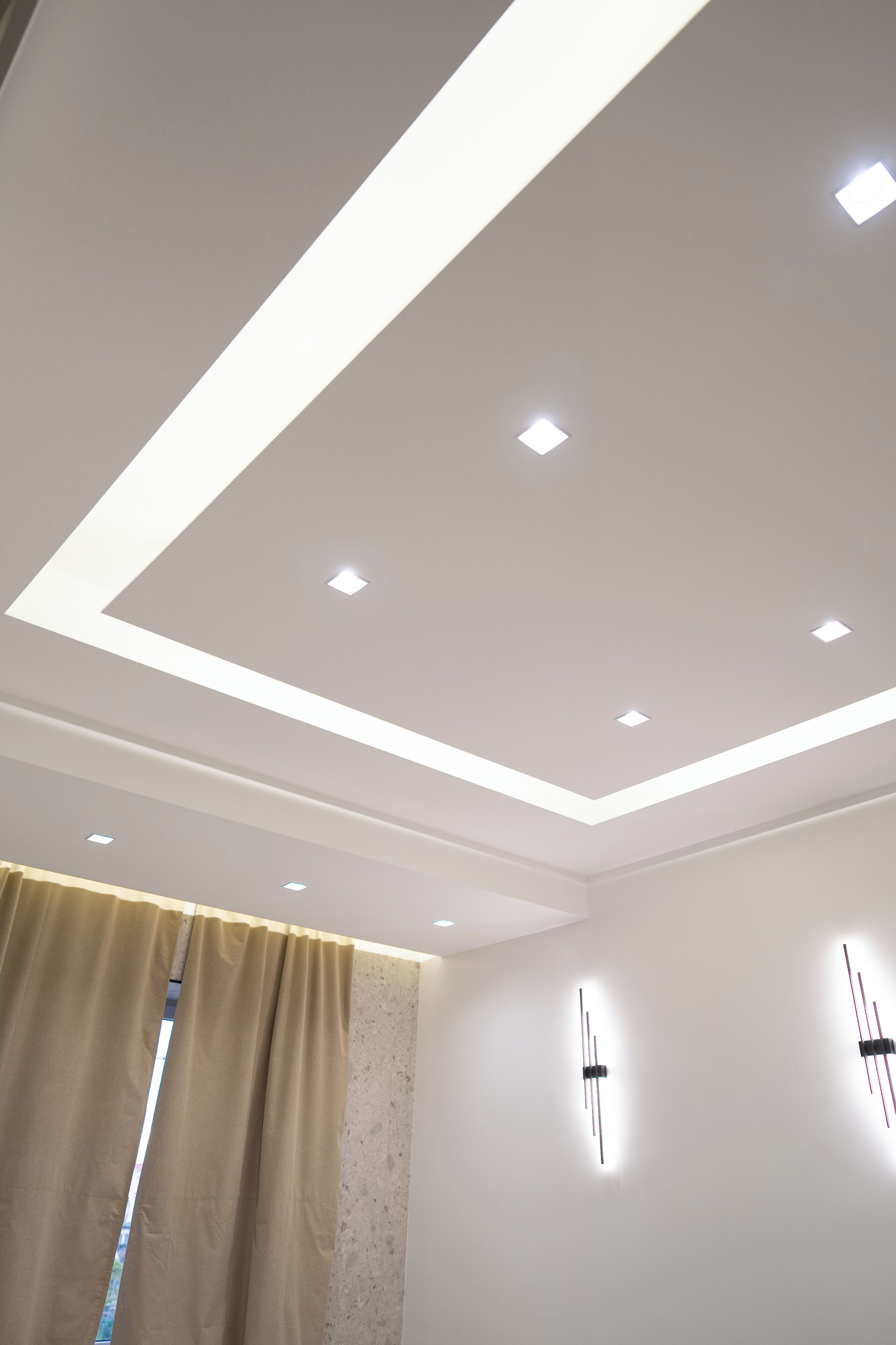 Une chambre moderne avec un plafond en retrait, une fenêtre avec des rideaux et des luminaires fixés au mur.