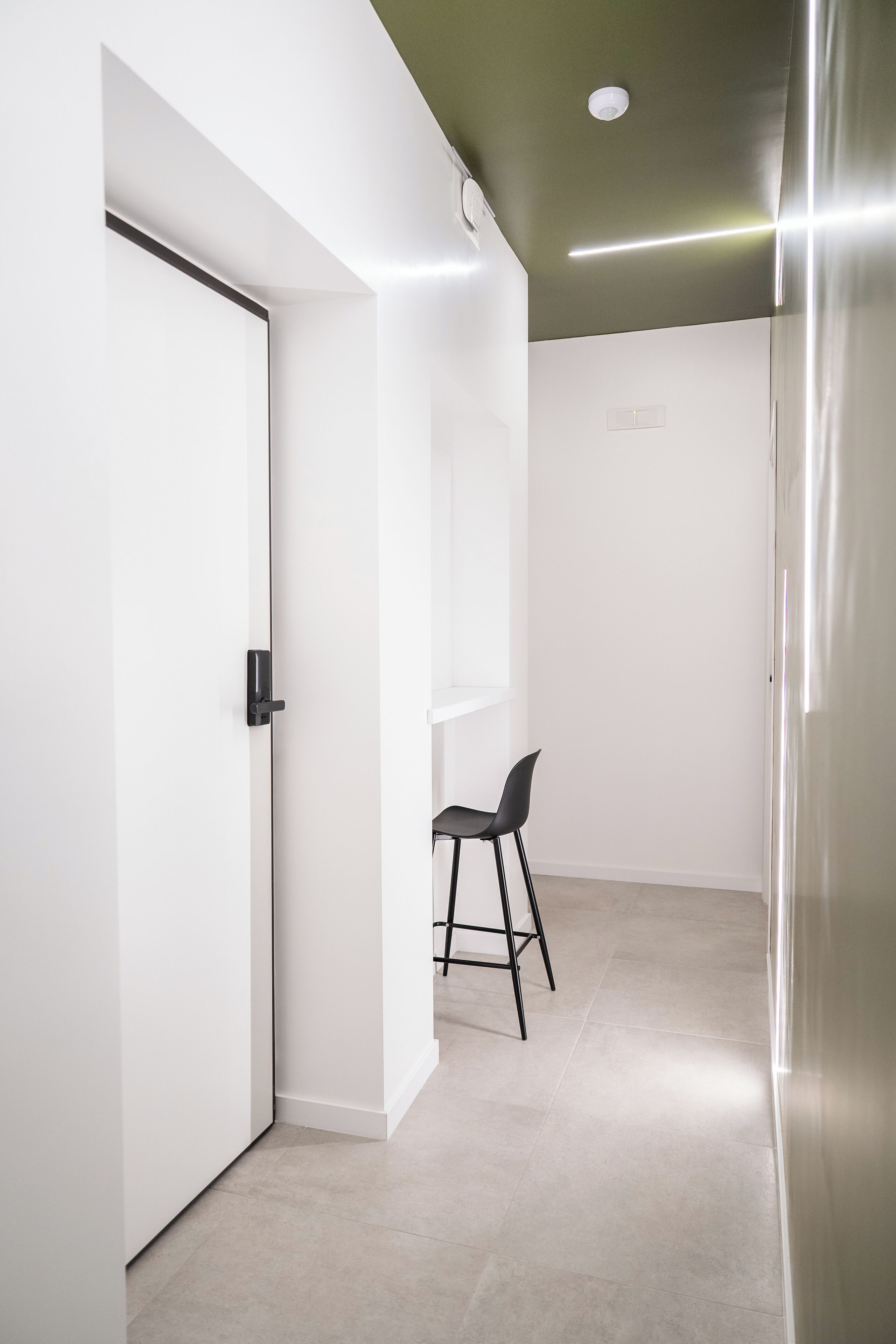 Um corredor moderno e minimalista com uma porta fechada, uma pequena área de estar com uma poltrona, e uma luminária no teto.