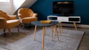 A modern living room with a sloped ceiling, a television on a white stand, two yellow armchairs, a round coffee table, and a grey rug. - Charming seaside apartment (Dinard)
