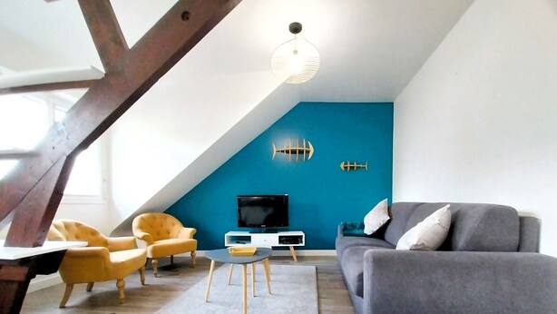A modern living room with a sloped ceiling, a grey sofa, yellow armchairs, a flat-screen TV, and decorative fish wall art. - Charming seaside apartment (Dinard)