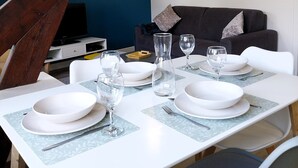 A dining area with a table set for four, white plates, glasses, and cutlery. A dark sofa is in the background, and a wooden beam structure is visible. - Charming seaside apartment (Dinard)
