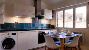 A modern kitchen with a built-in washing machine, a dining area with a table set for two, and large windows offering a view of the city. - Charming seaside apartment (Dinard)