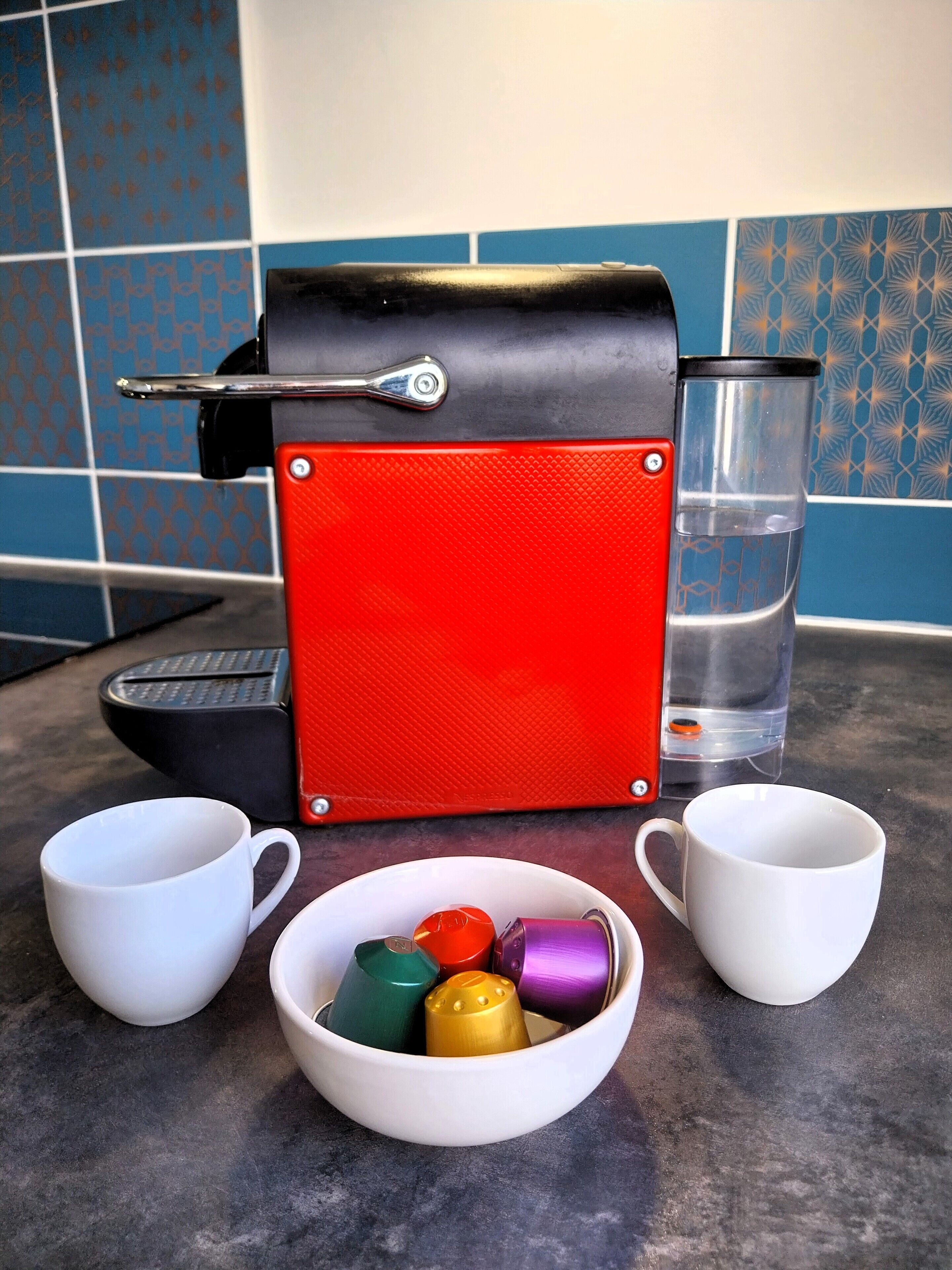 Un coin cuisine comprenant une machine à café, deux tasses blanches et un bol rempli de bouchons de bouteille de différentes couleurs.