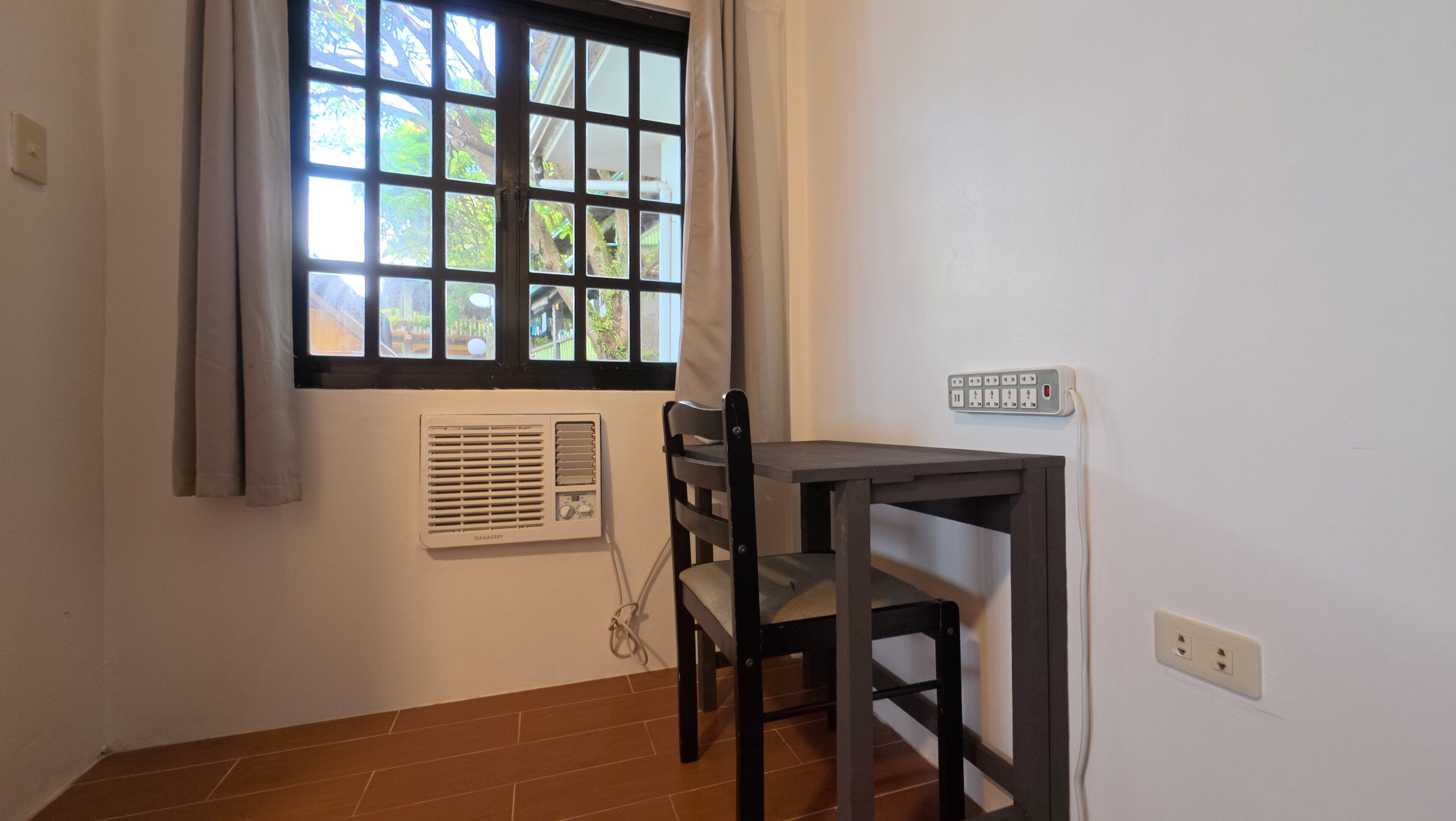 Una habitación pequeña con ventana, una mesa, una silla, un enchufe y aire acondicionado instalado en la pared.