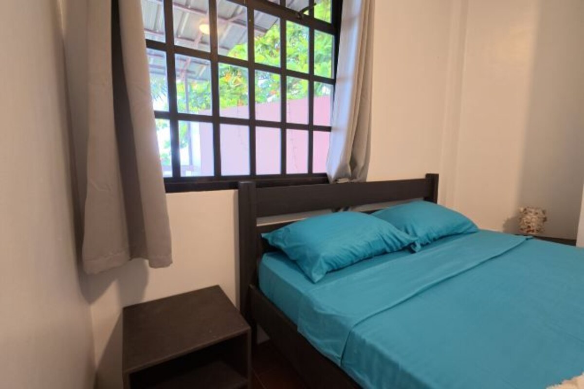 A room with a bed, a nightstand, a window with curtains, and a small table with a vase.