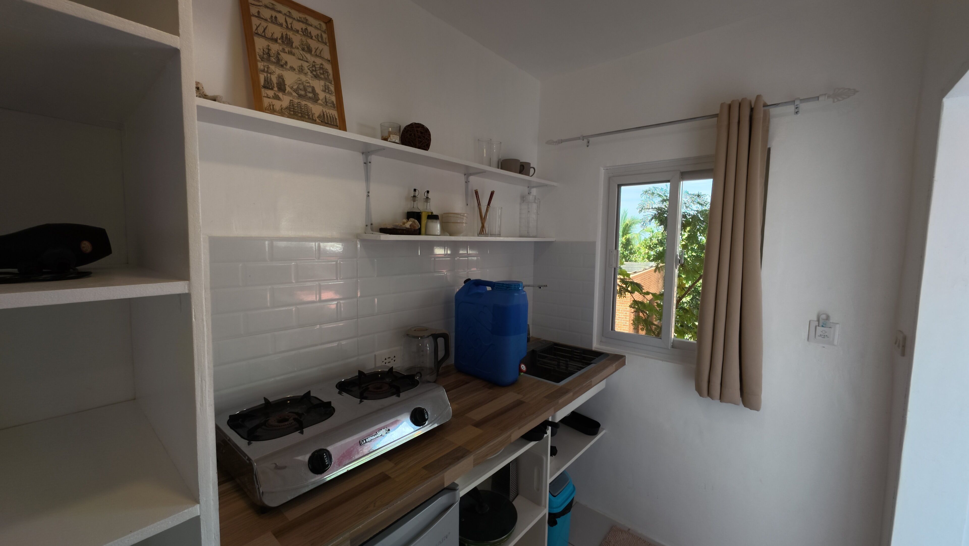 A compact kitchen with white cabinets, a gas stove, a blue water container, and a window with curtains.