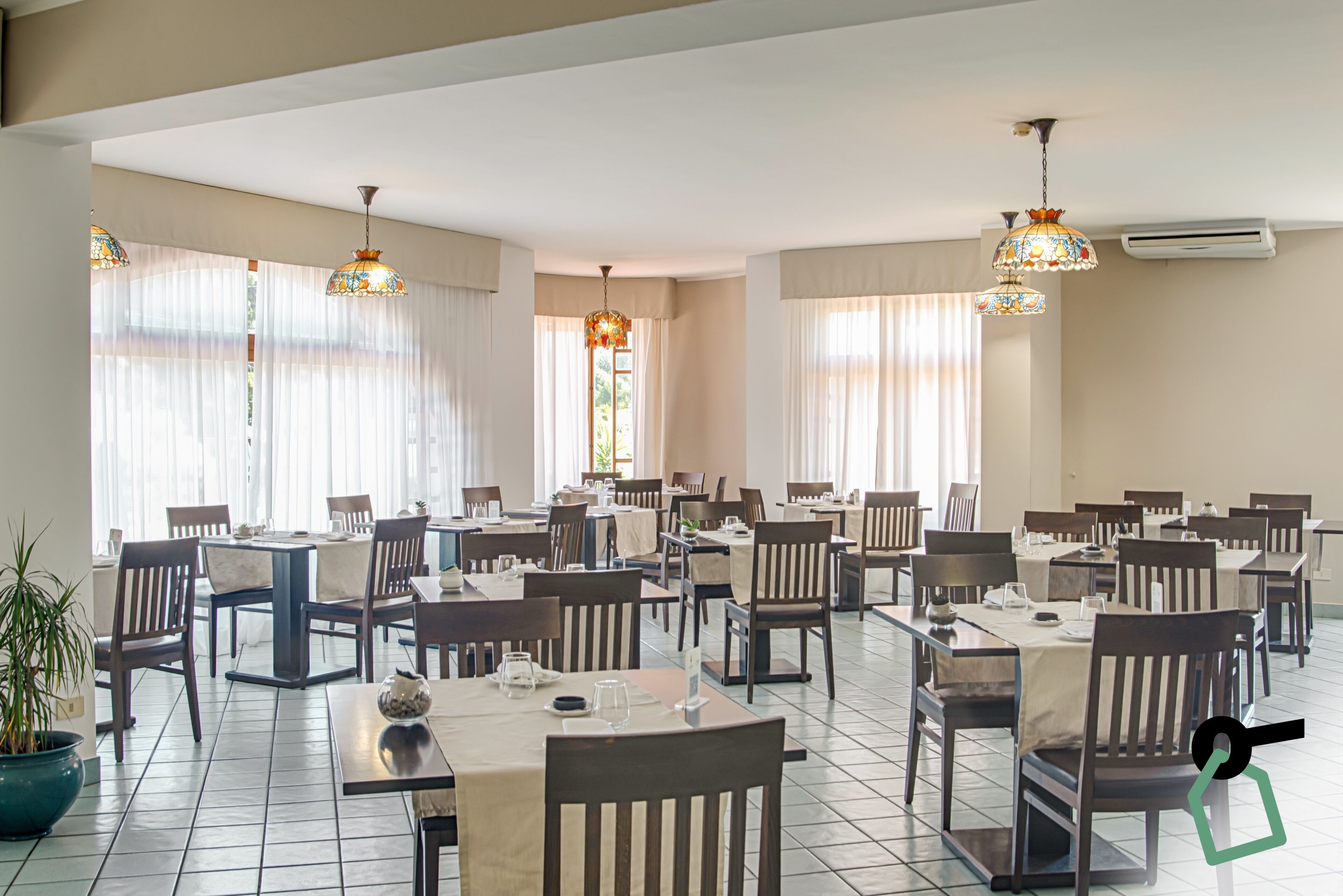 Un vaste espace dédié à la salle à manger, avec de nombreuses tables et chaises, de grandes fenêtres ornées de rideaux légers et des lampes décoratives suspendues au plafond.
