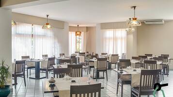 Un vaste espace dédié à la salle à manger, avec de nombreuses tables et chaises, de grandes fenêtres ornées de rideaux légers et des lampes décoratives suspendues au plafond.