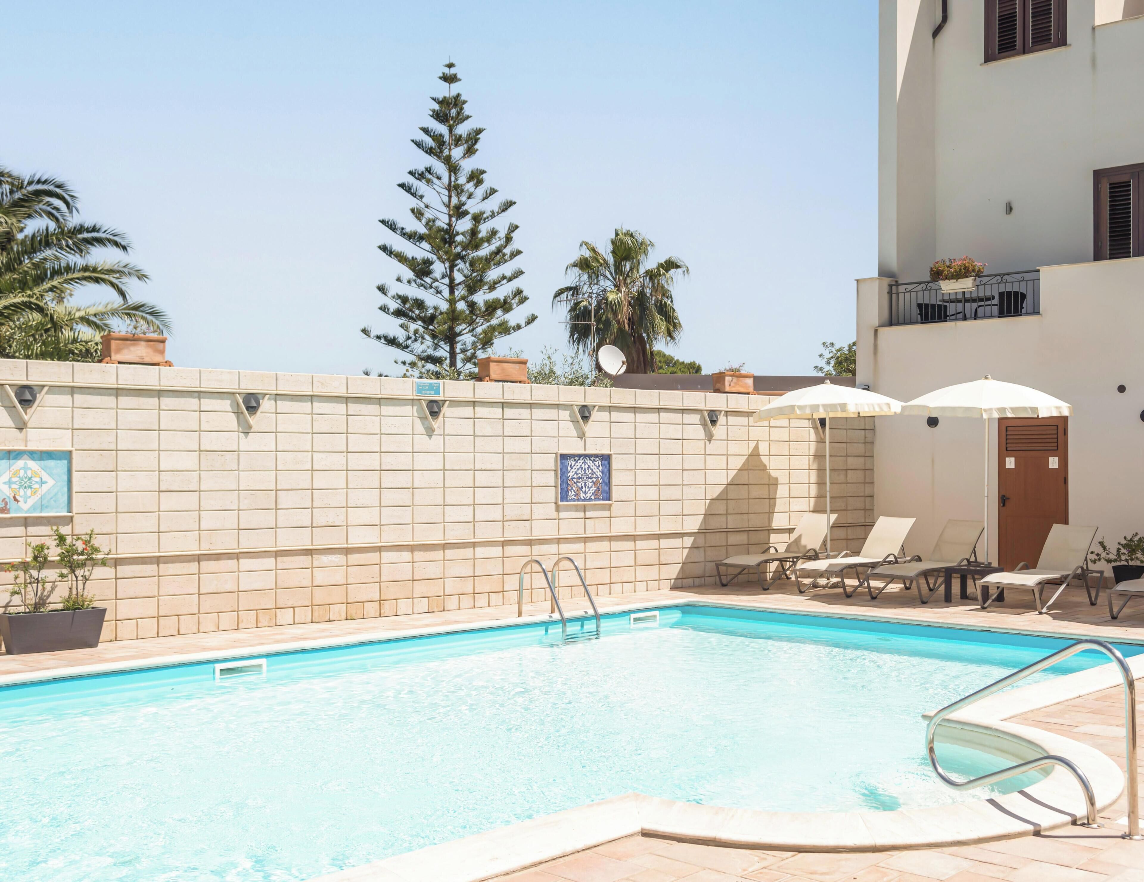 A swimming pool with lounge chairs and umbrellas, surrounded by a tiled wall with decorative tiles and potted plants.