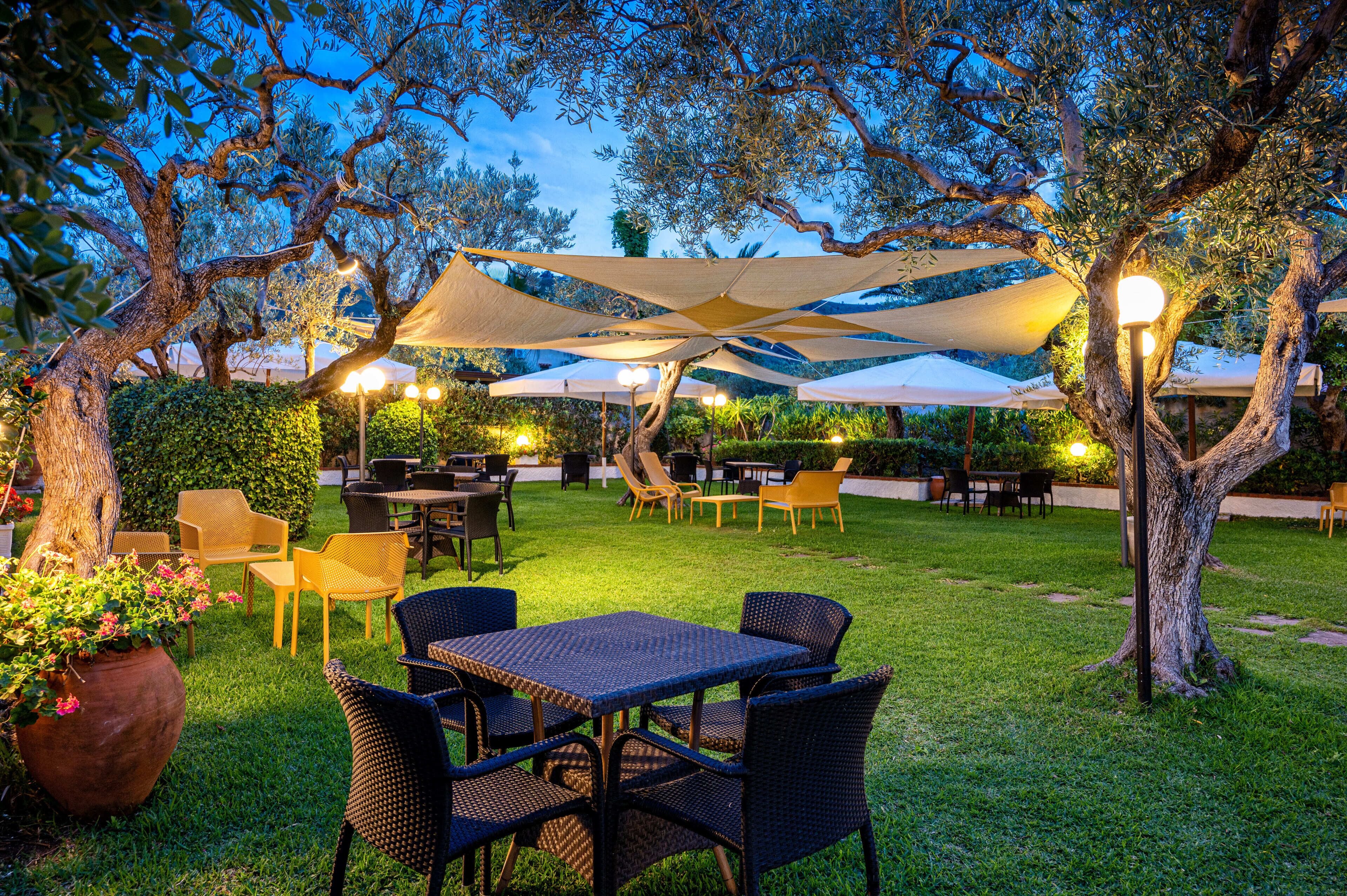 An outdoor patio area with tables and chairs, shaded by large umbrellas, surrounded by trees and greenery.