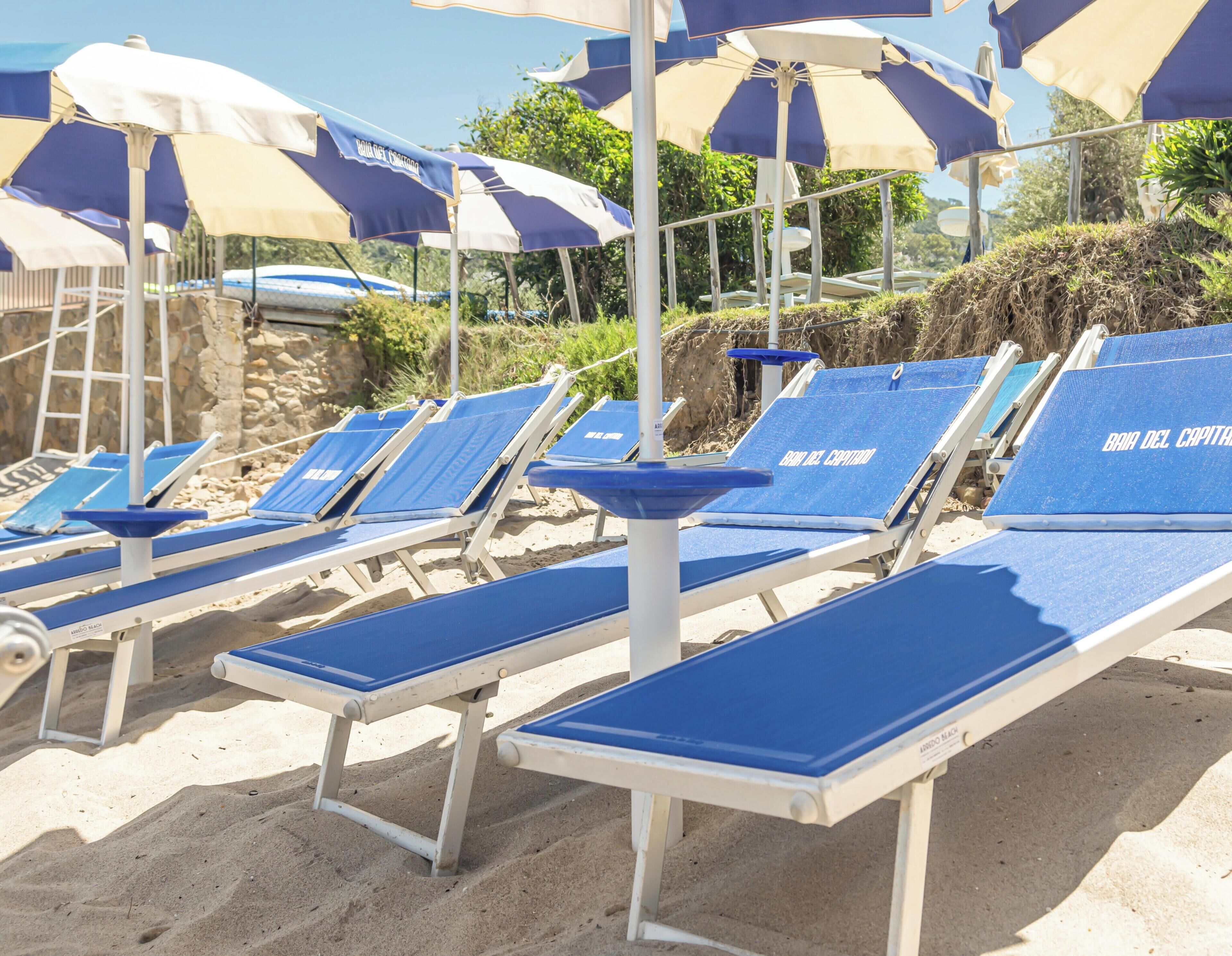 Eine Reihe aus Liegestühlen mit blauen Kissen und weißen Gestellen, angeordnet auf einem Sandstrand, der mit blau-weißen Strandsonnenschirmen beschattet wird.
