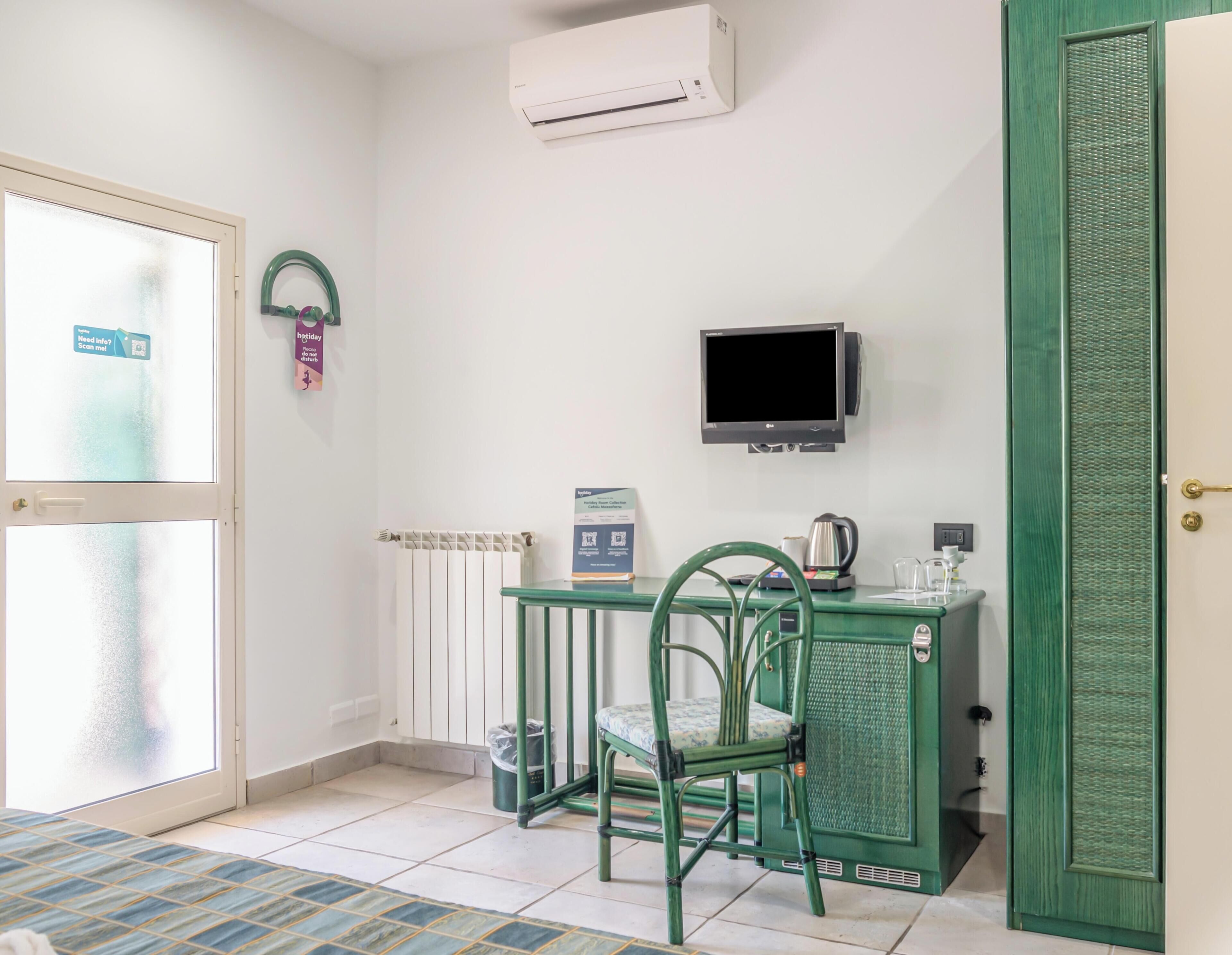 Una stanza piccola con una poltrona verde, un tavolo con un bollitore, una TV e una porta con maniglia.