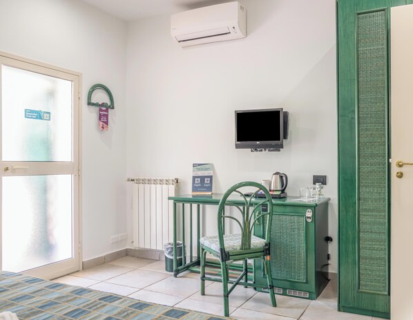 Une petite pièce avec une chaise verte, une table sur laquelle se trouve une bouilloire, un téléviseur et une porte dotée d’une poignée.