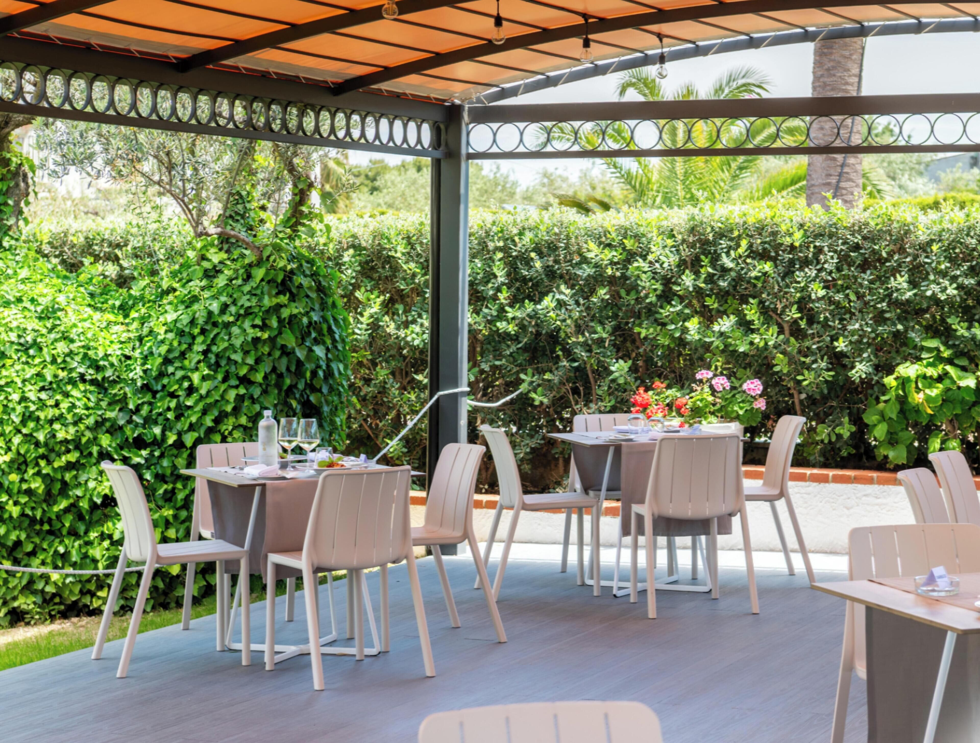 Un patio al aire libre con mesas y sillas, rodeado de exuberante follaje y una estructura de pérgola.