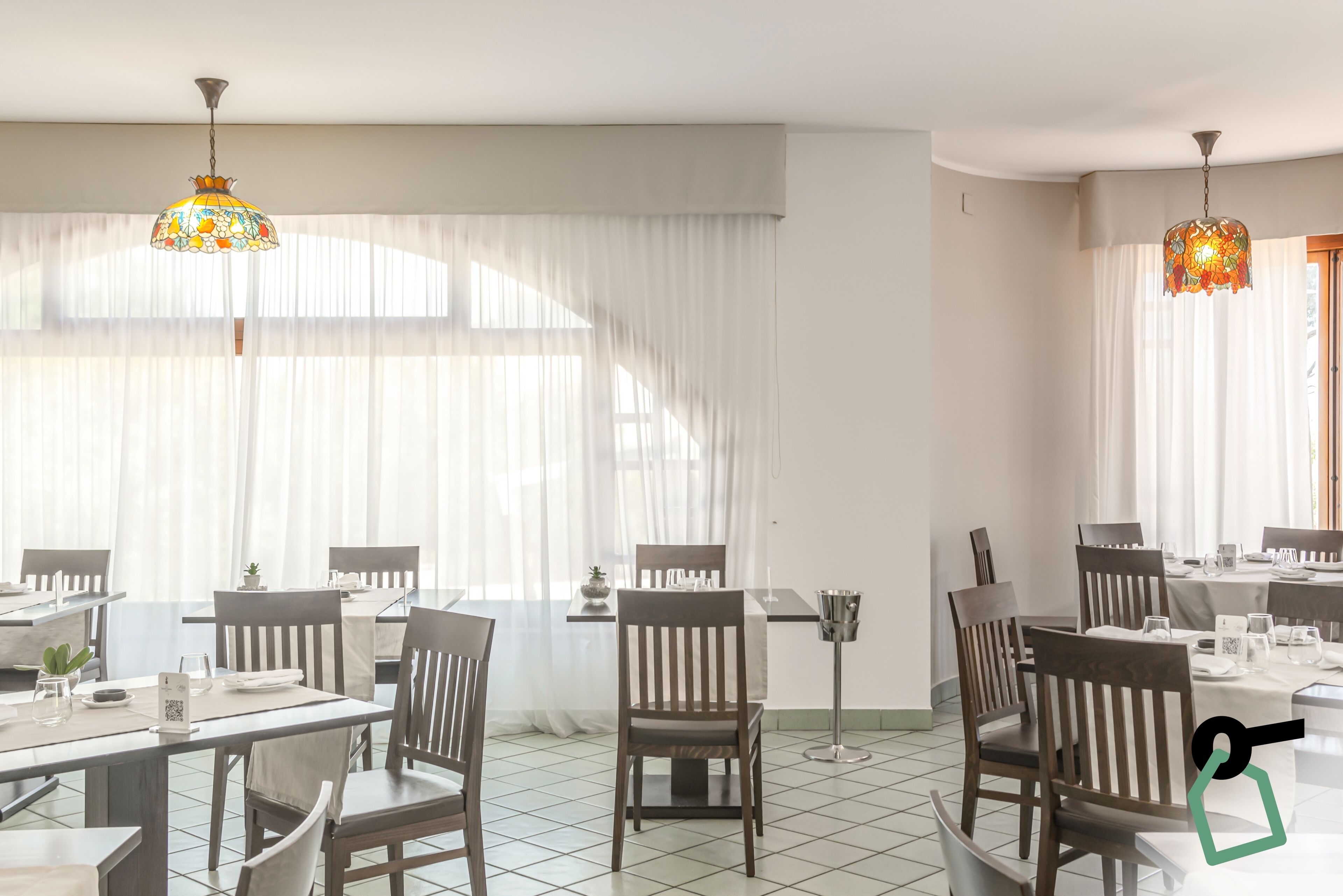 Een eetruimte met tafels en stoelen, grote ramen voorzien van dunne gordijnen en decoratieve hanglampen.