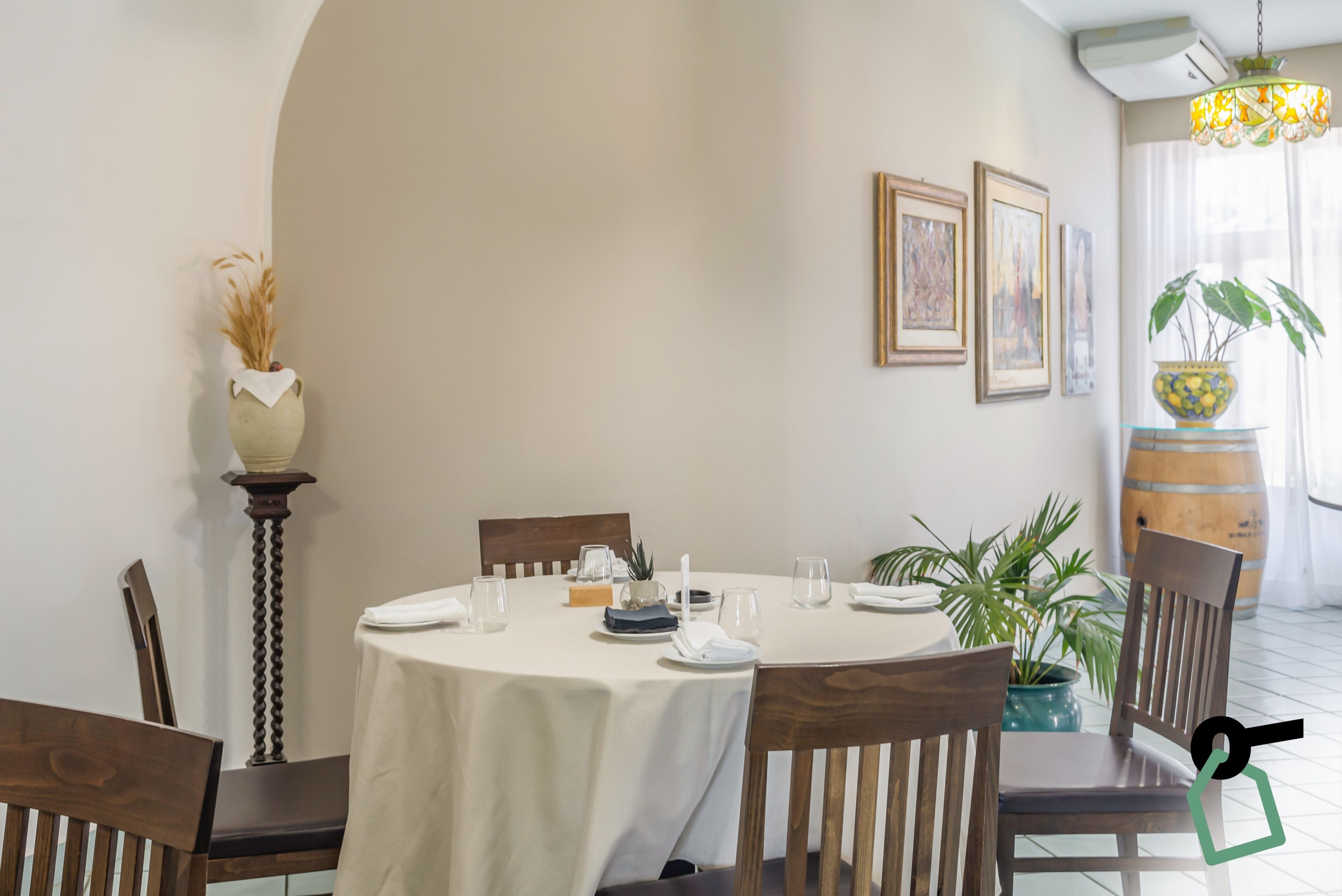 Un coin repas avec une table ronde dressée pour quatre personnes, entourée de chaises en bois. Des œuvres d’art encadrées ornent le mur, ainsi qu’une plante en pot et un vase décoratif contenant des plantes séchées.