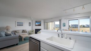 A modern kitchen with a large white countertop, a double sink, and a dishwasher. There is a dining area with a gray sofa and a small table. The room has large windows with a view of buildings outside.