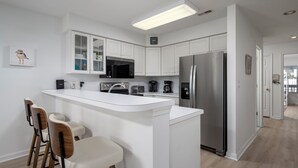 A modern kitchen with white cabinetry, a stainless steel refrigerator, a microwave, and a coffee maker. There are three bar stools and a large island with a sink.