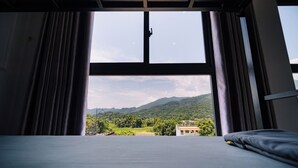 A room with a bed facing a large window, curtains, and a view of a green landscape with mountains. - Ha Giang City Hostel (Ha Giang 1)