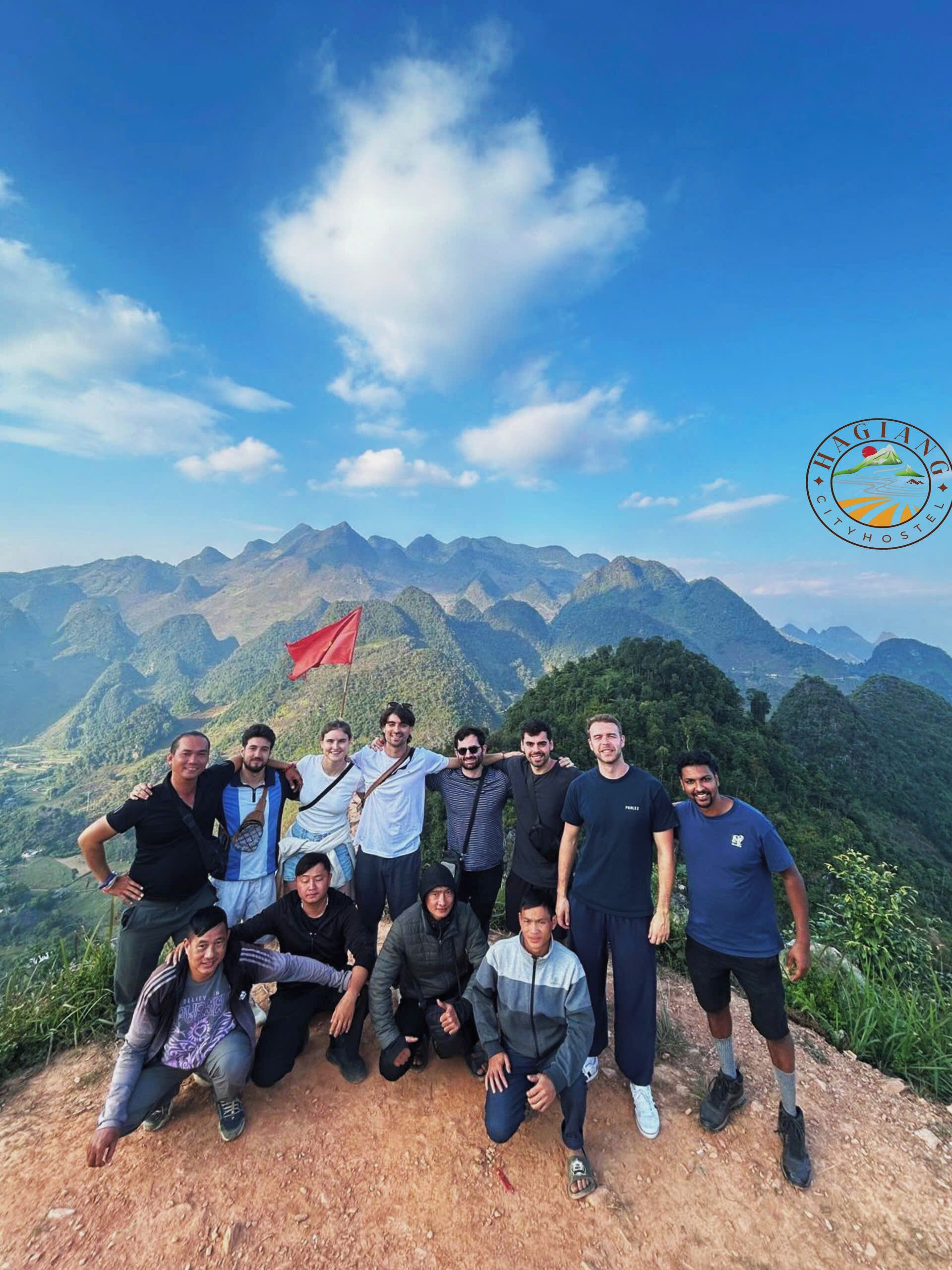 Um grupo de pessoas posando em um cume de montanha com uma bandeira vermelha, com vista para um vale e montanhas ao fundo.