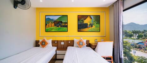 Habitación de hotel con dos camas individuales, un cabecero de madera, una mesita de noche, un escritorio y un ventanal con vistas a un paisaje urbano y montañas.