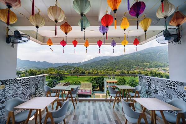 A balcony with tables and chairs, a sign for a fresh pizza pasta restaurant, and a view of green mountains. - Ha Giang City Hostel (Ha Giang 1)