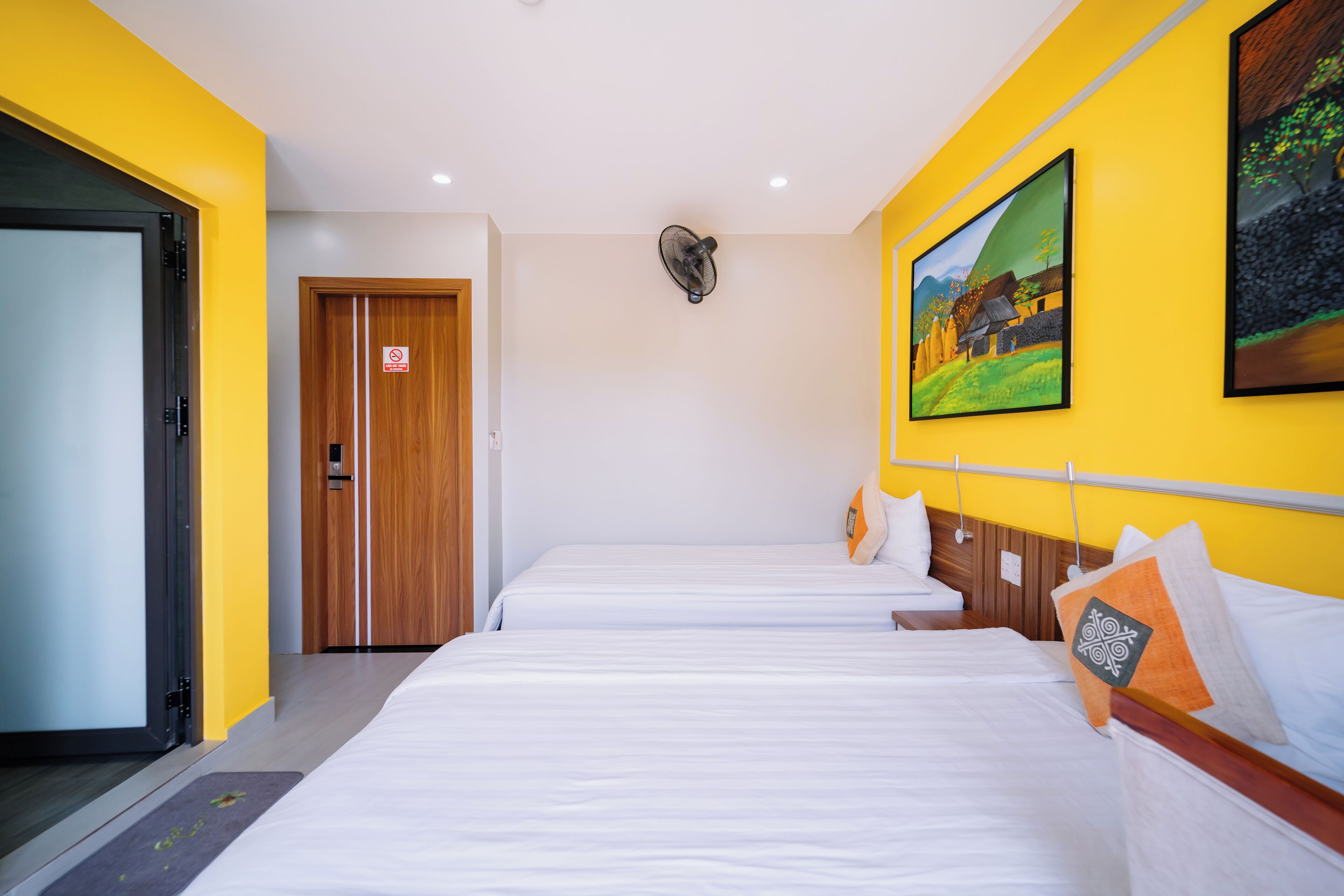 A hotel room with two beds, a wooden door, a fan, and a painting on the wall.