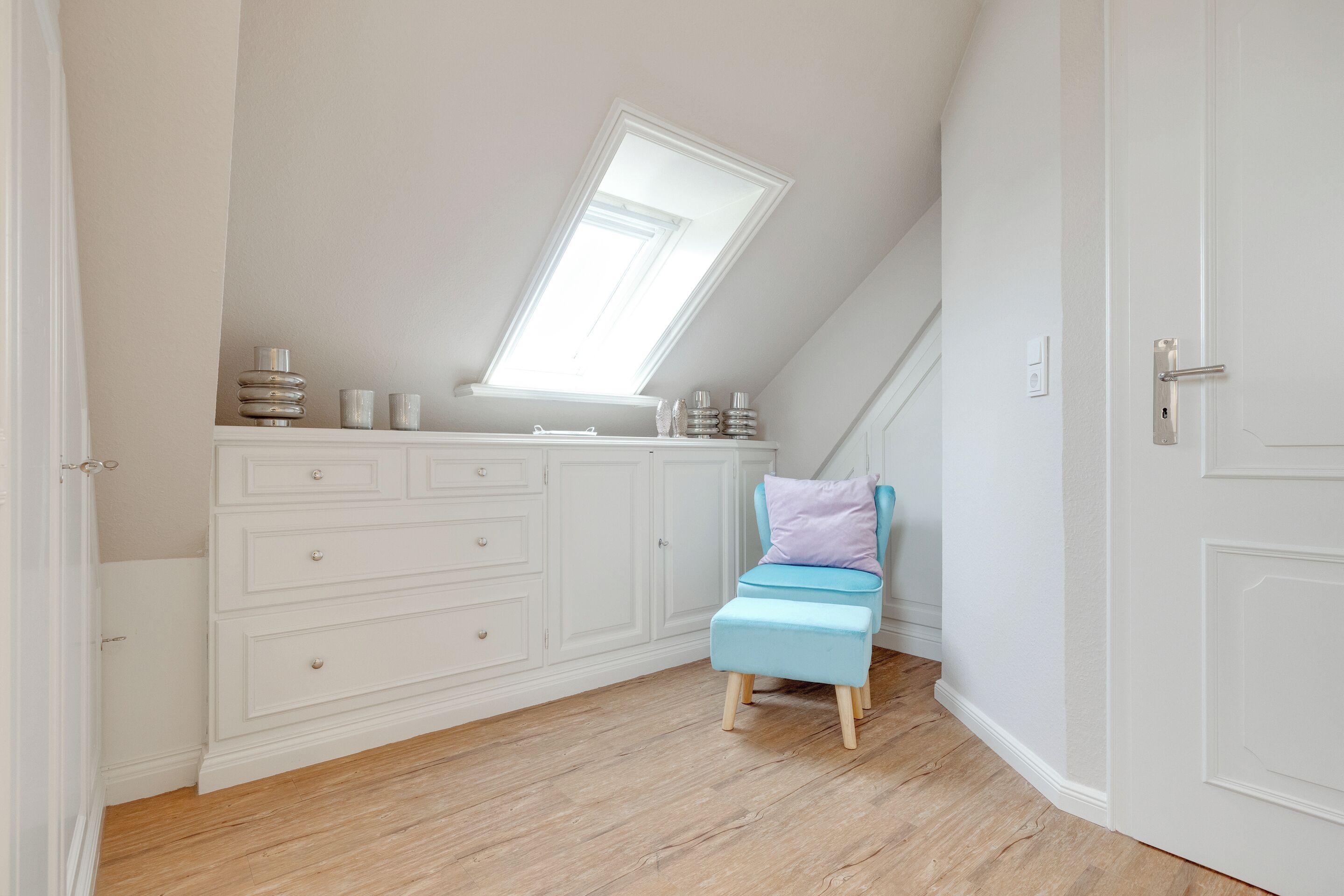 Une pièce avec un plafond en pente, un puits de lumière, un meuble blanc avec des tiroirs, une petite chaise bleue avec un coussin et un plancher en bois.