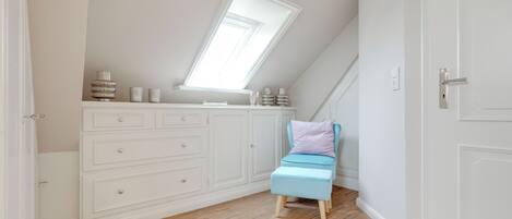 Une pièce avec un plafond en pente, un puits de lumière, un meuble blanc avec des tiroirs, une petite chaise bleue avec un coussin et un plancher en bois.