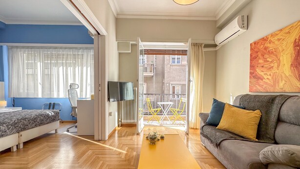 Una sala de estar moderna con un sofá, una mesa de centro y un televisor. Hay un balcón con vista a edificios.