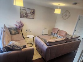 Un soggiorno con un divano beige, un piccolo tavolino rotondo e due poltrone. Si vede un termosifone, un lampadario a parete e un elemento decorativo a parete.