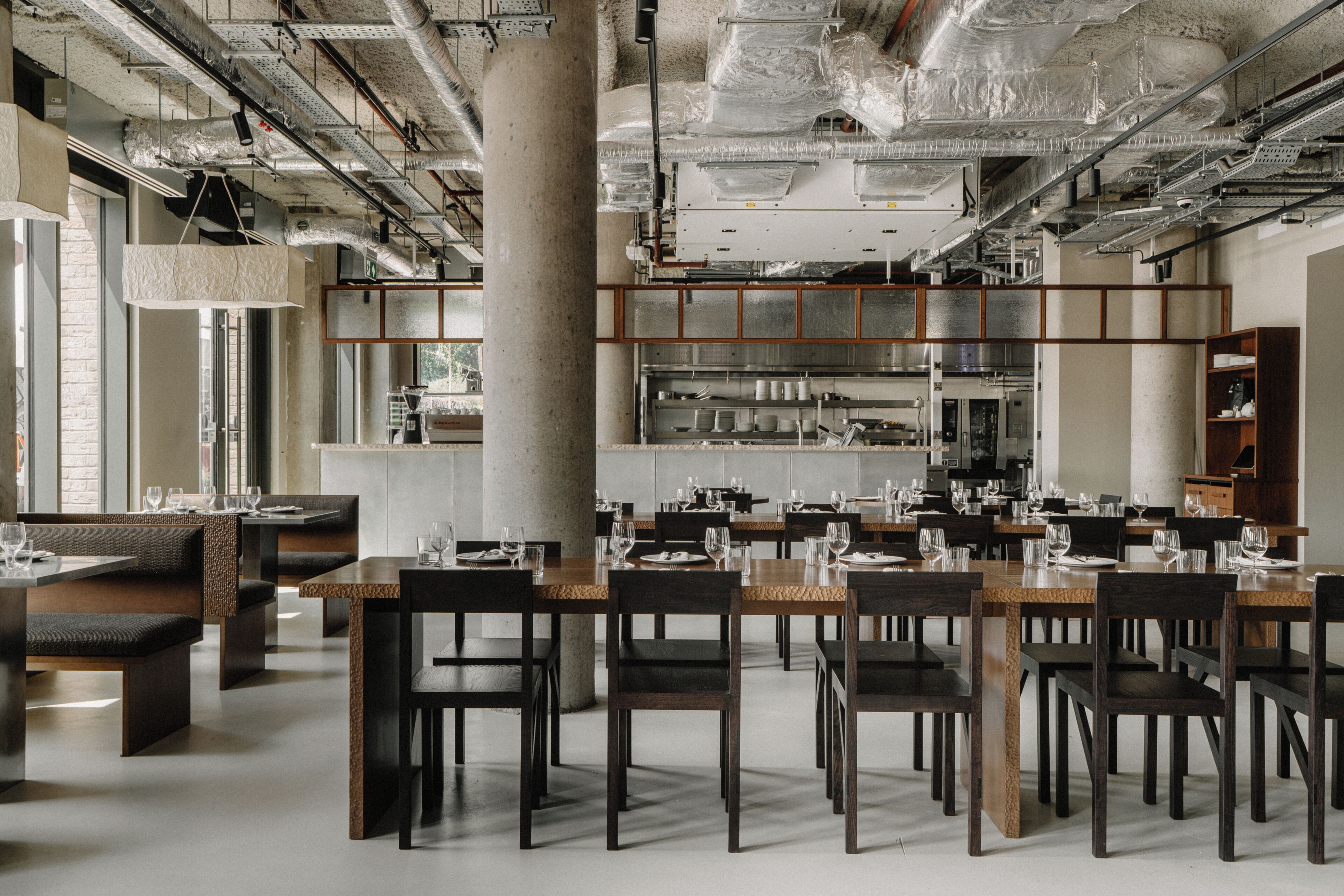 Un restaurant moderne avec de hauts plafonds, des conduits apparents et une cuisine centrale. Les tables sont dressées avec des nappes blanches et des verres à vin.