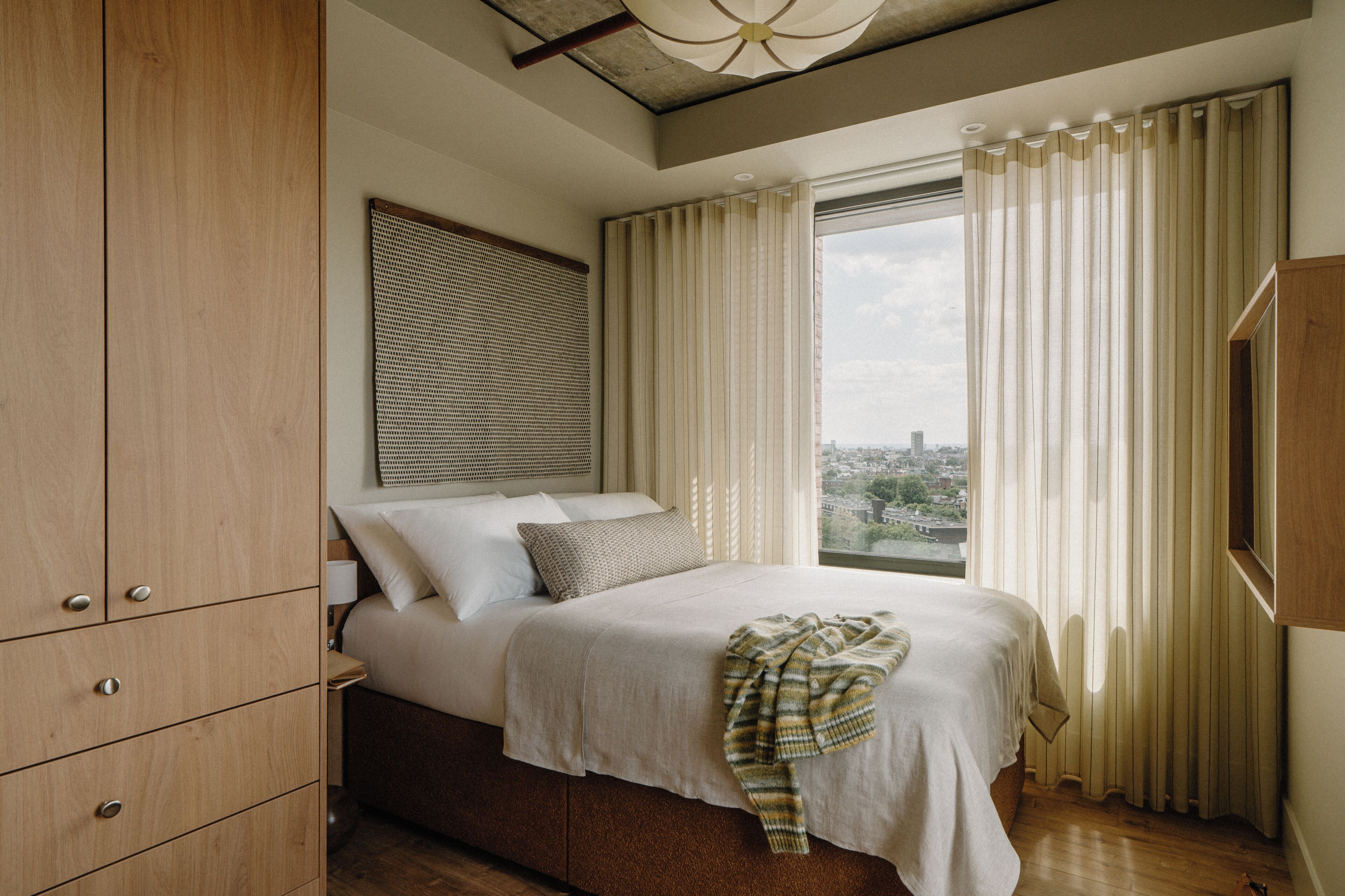 A bedroom with a bed, wooden wardrobe, and a city view through the window.
