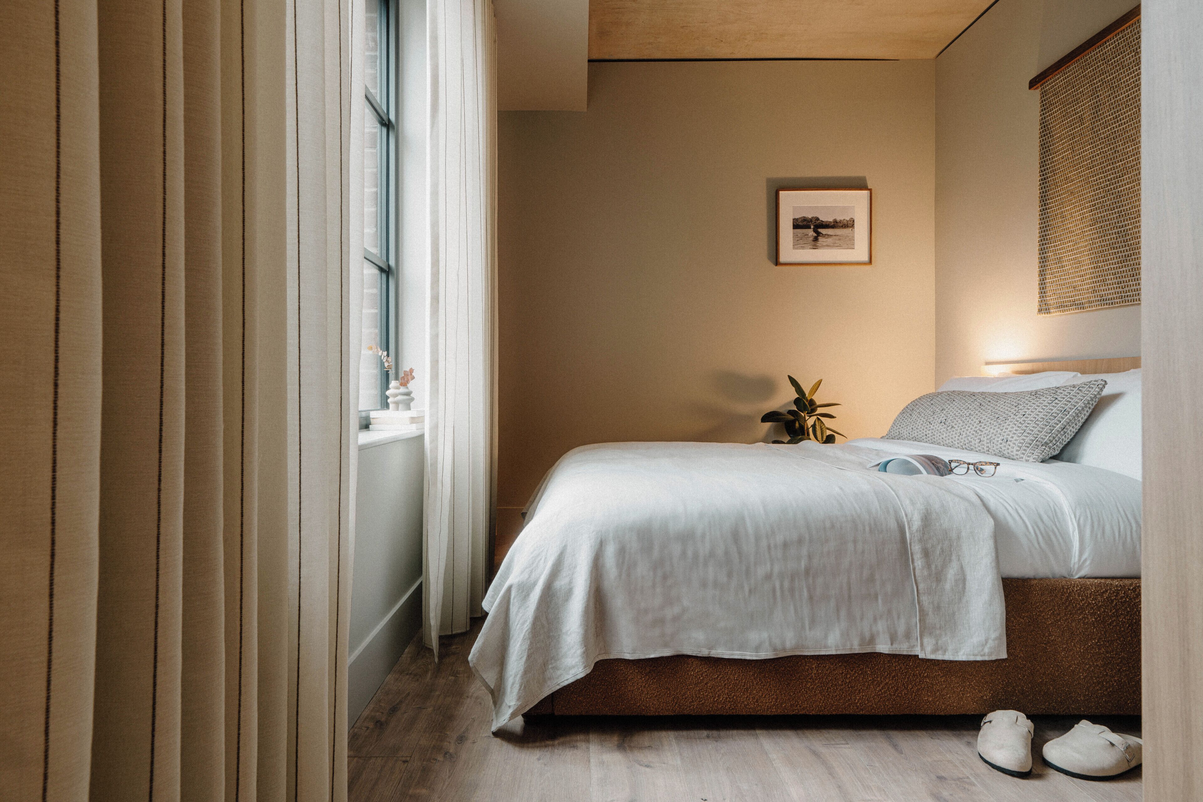 A bedroom with a bed, a window with curtains, a framed picture on the wall, and a pair of shoes on the floor.