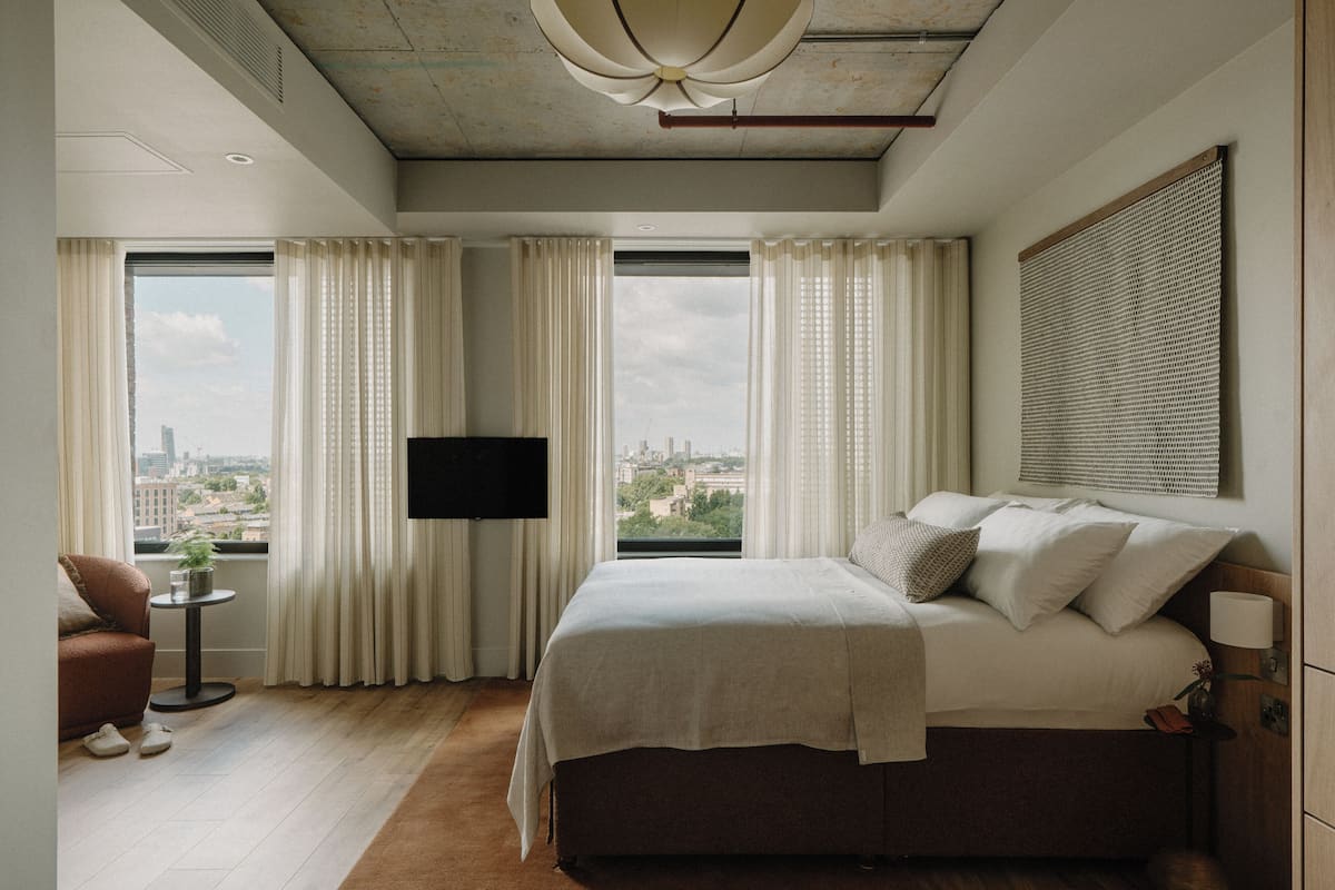 A modern bedroom with a large bed, a TV mounted on the wall, and a city view through the windows.