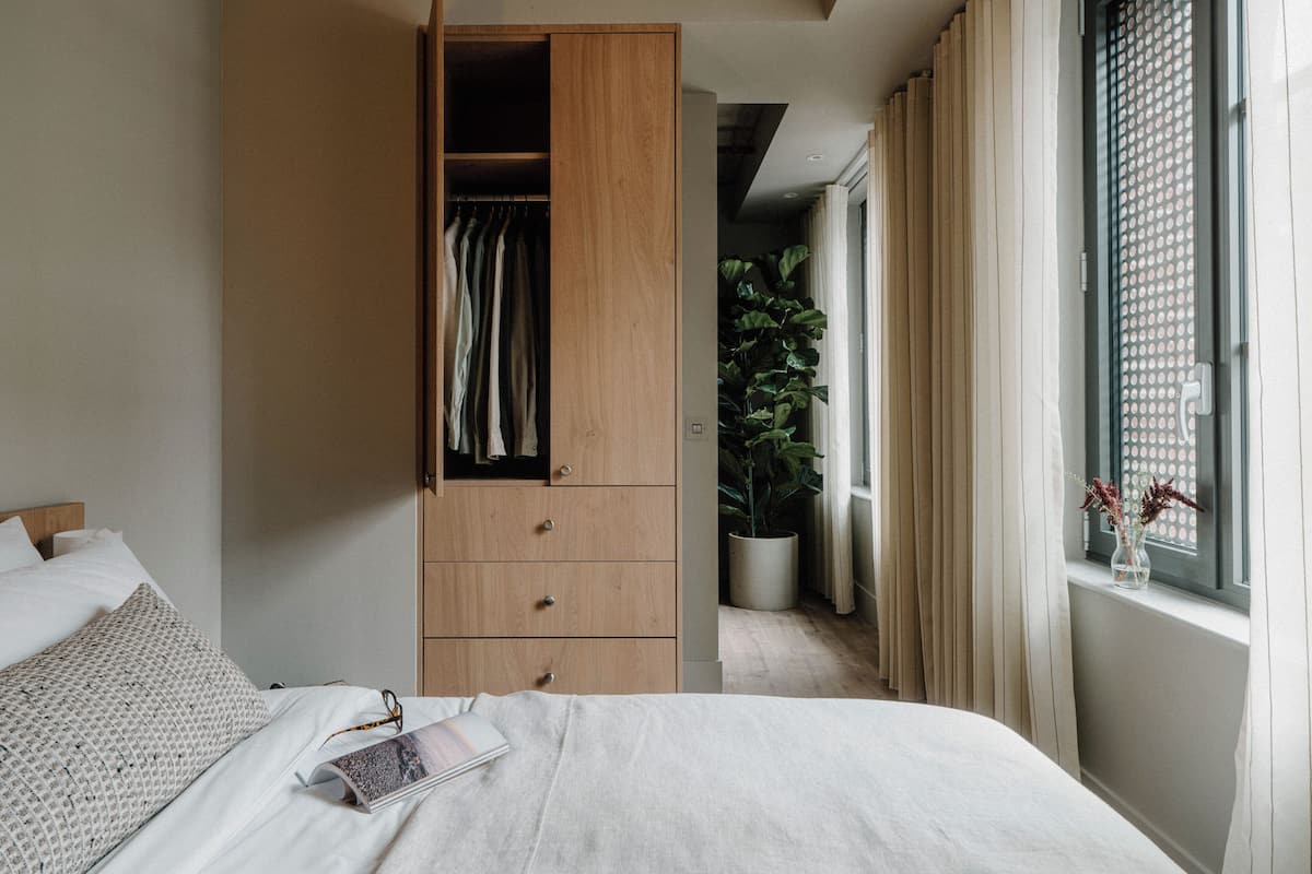 A bedroom with a bed, a wooden wardrobe, a potted plant, and a window with curtains.