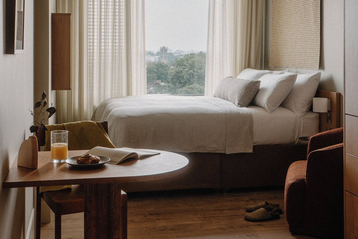 A hotel room with a bed, a dining table with a chair, a lamp, and a view of the city through the window.