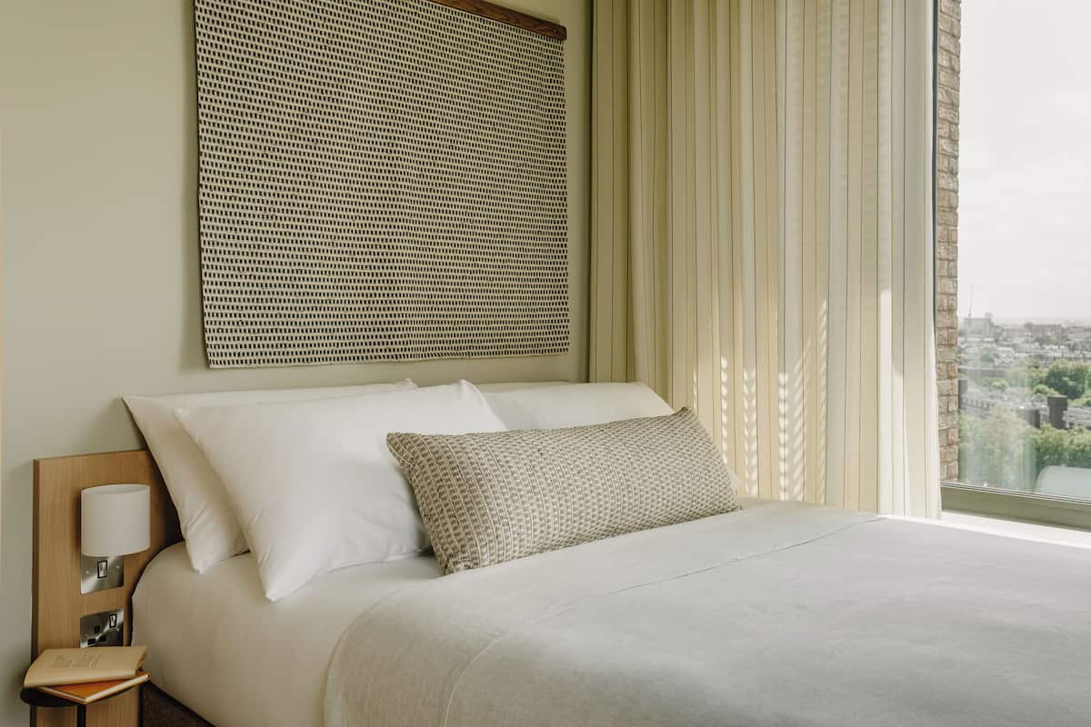 A neatly made bed with a headboard, a bedside table with a lamp, and a view of a cityscape through a window with curtains.