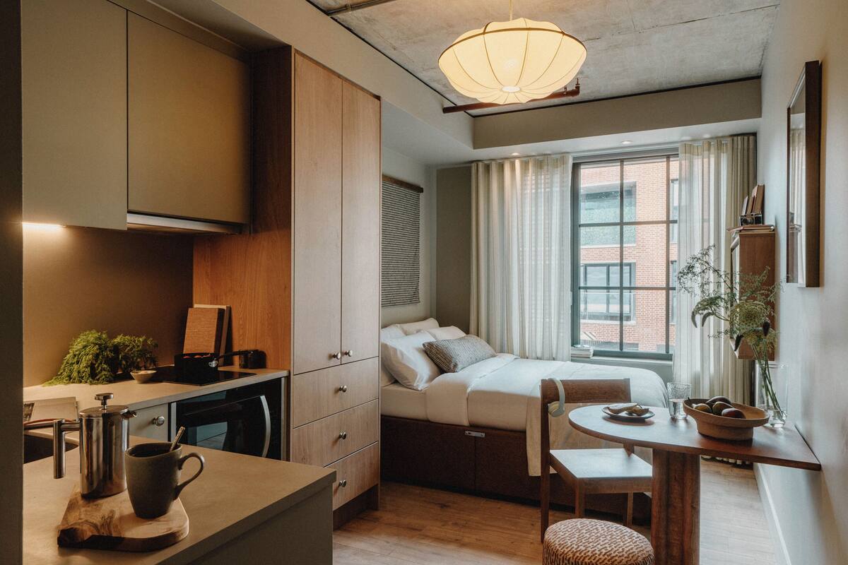 A compact hotel room with a kitchenette, a bed, a dining area, and a large window with curtains.
